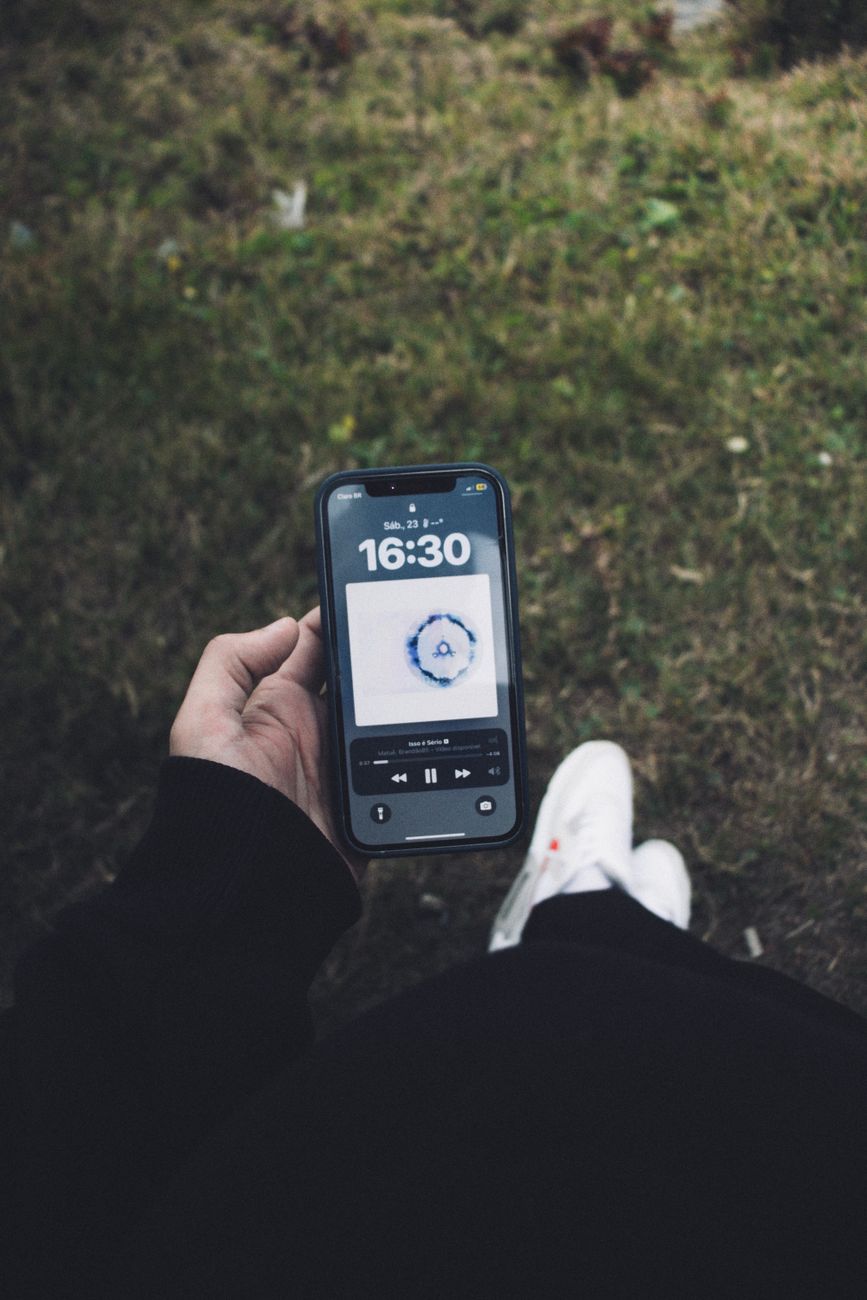 A person holds a smartphone showing a music app and the time 16:30 outdoors.
