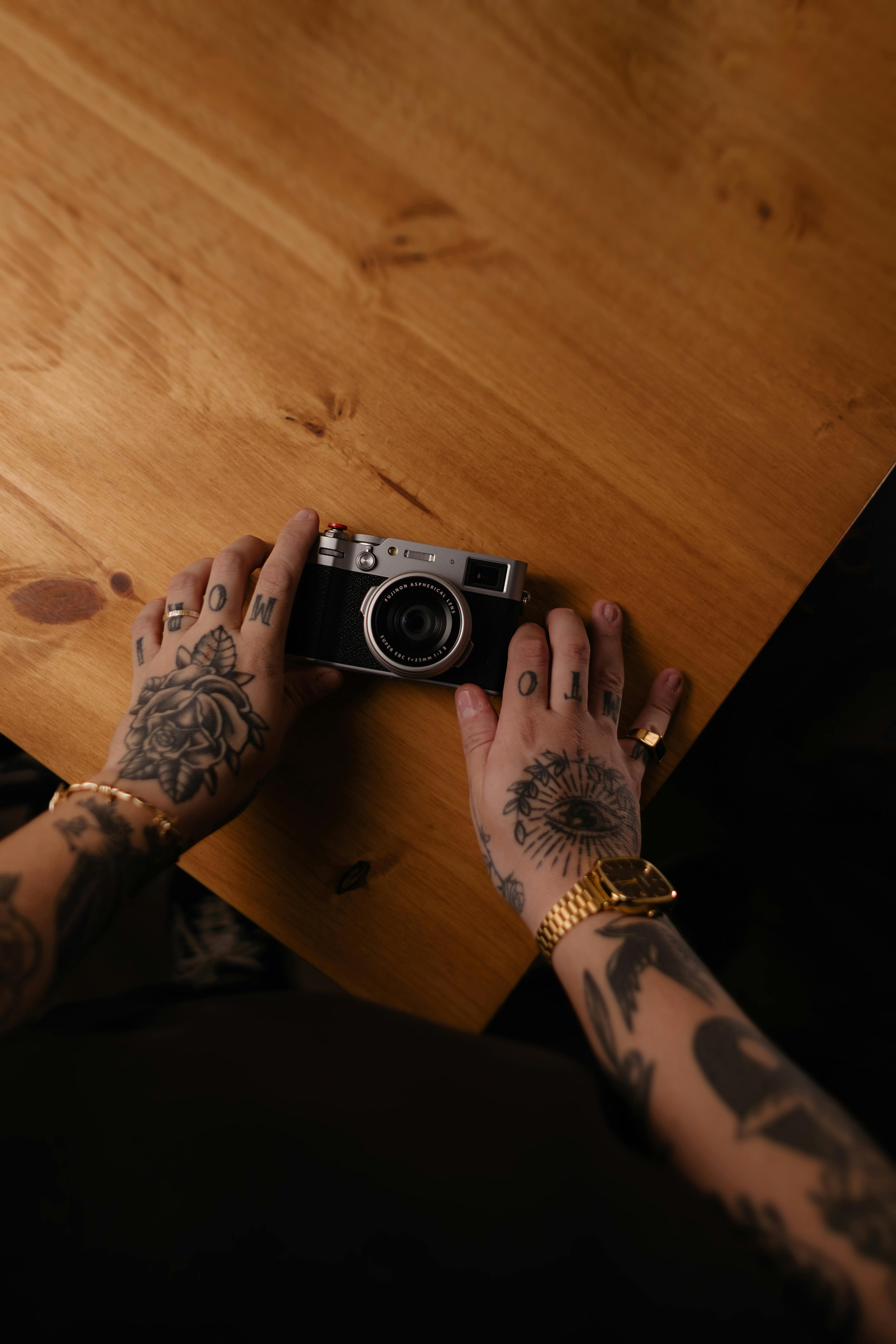 Gratis Oppdag kunsten å fotografere med tatoverte hender som holder et klassisk kamera på et rustikt trebord. Arkivbilde