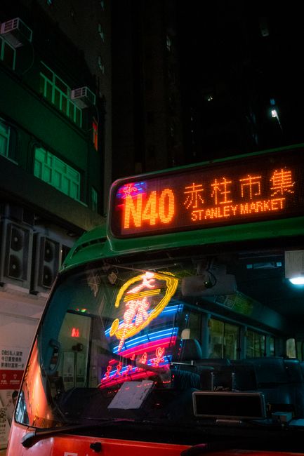 A vibrant night scene of a bus heading towards Stanley Market, illuminated by neon lights.