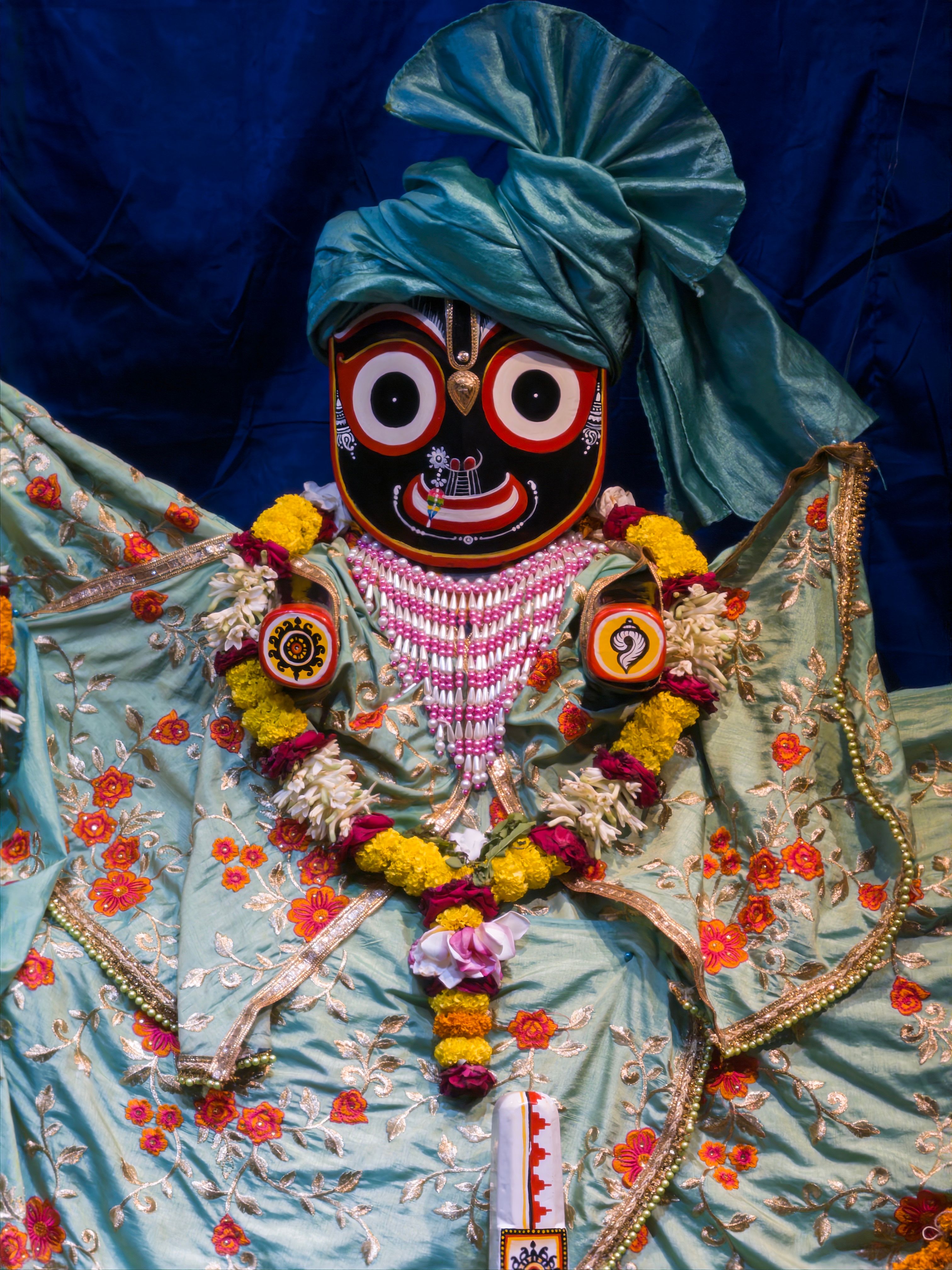 A vibrant Jagannath idol adorned in colorful floral garments and a turban, surrounded by garlands.