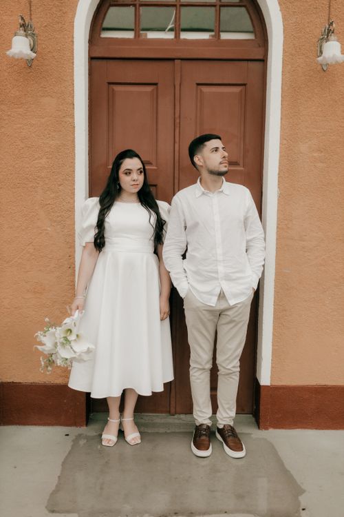 Free Elegant couple in white attire posing against a rustic brown doorway, embracing a timeless wedding theme. Stock Photo