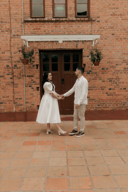 Free Couple in white attire posing outdoors against a rustic brick building backdrop, creating an elegant scene. Stock Photo