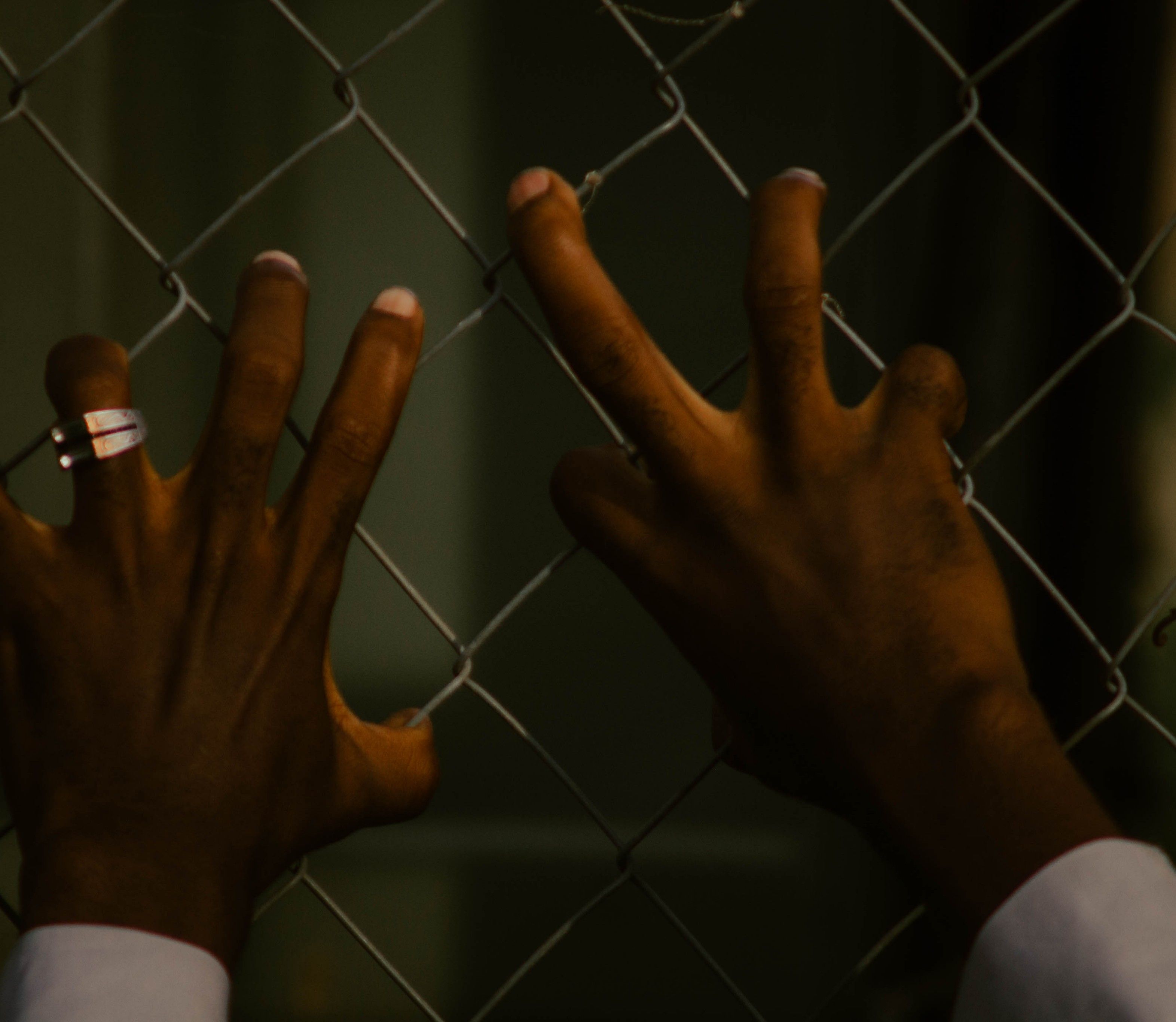 Close-up of hands gripping a metal fence, evoking themes of confinement and longing.