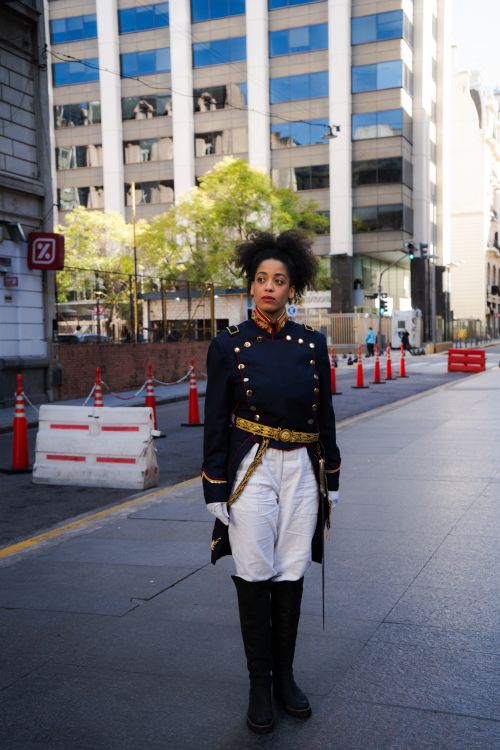 Free A woman in a historical military costume stands on a city street, surrounded by tall buildings and traffic cones. Stock Photo