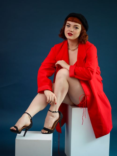 Free Stylish woman sits confidently in a red coat with black heels against a blue background. Stock Photo