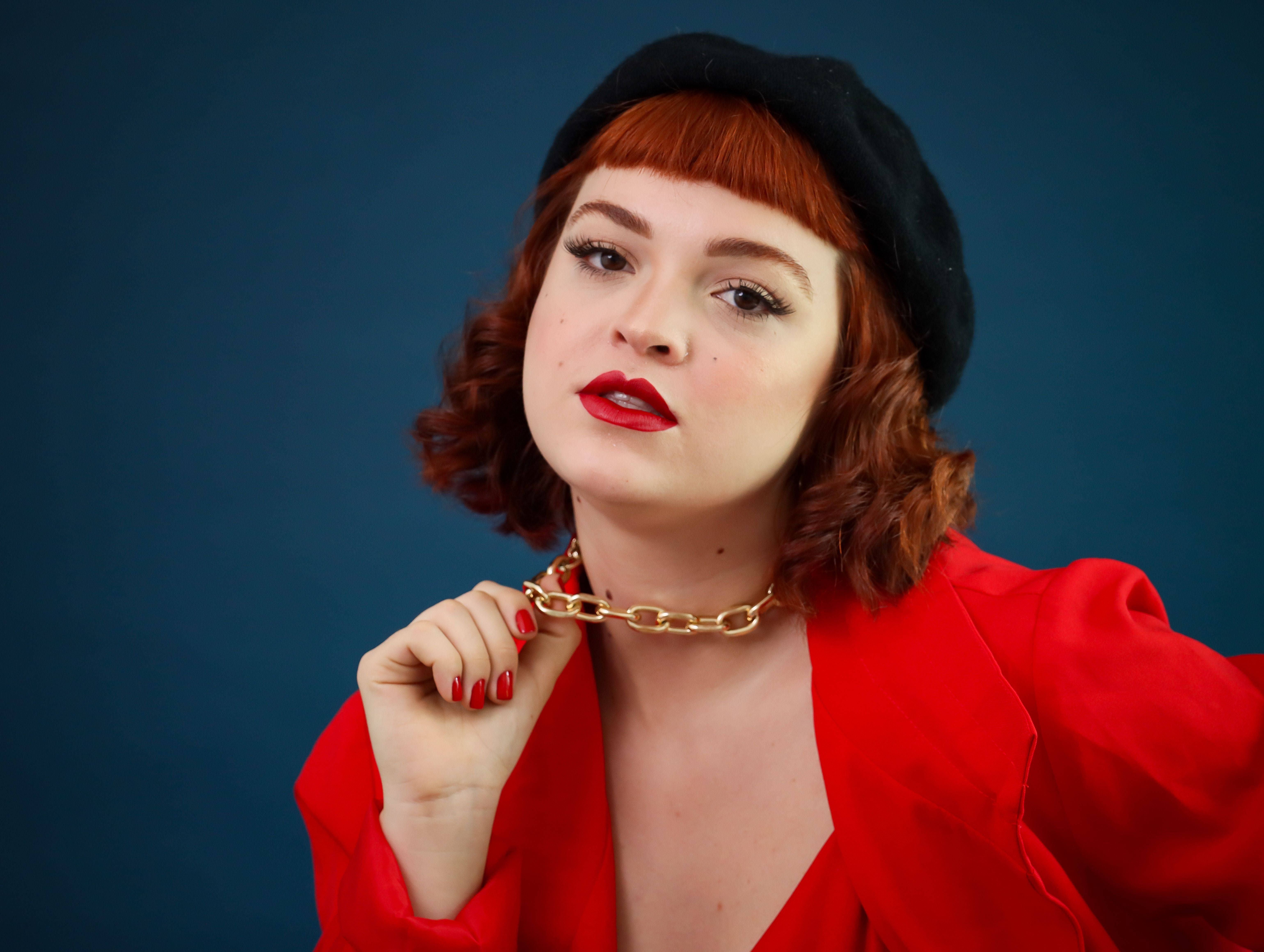 Fashionable woman posing confidently in red attire with bold chain jewelry against blue background.