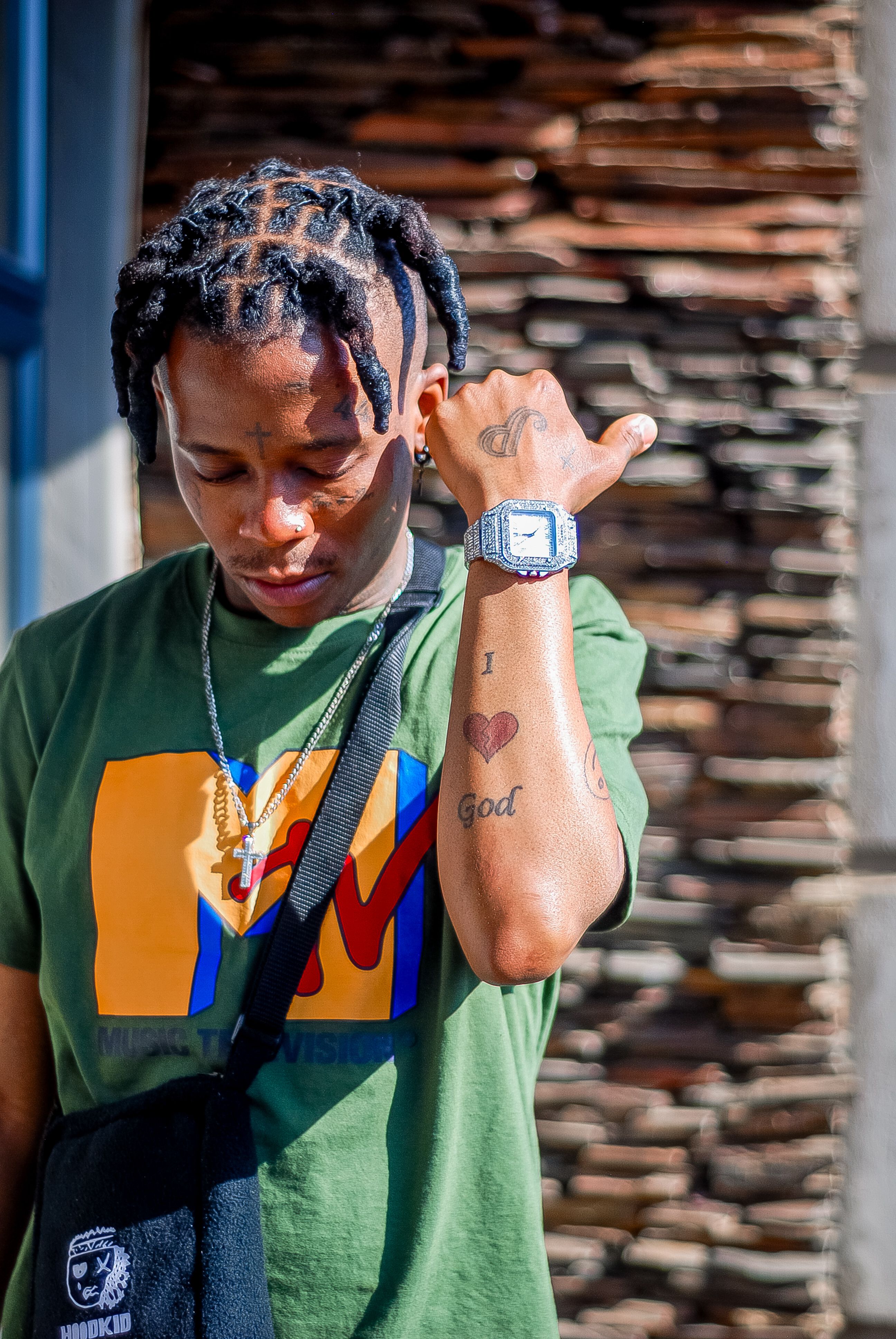 A young man with tattoos and trendy attire posing outdoors against a stone wall.