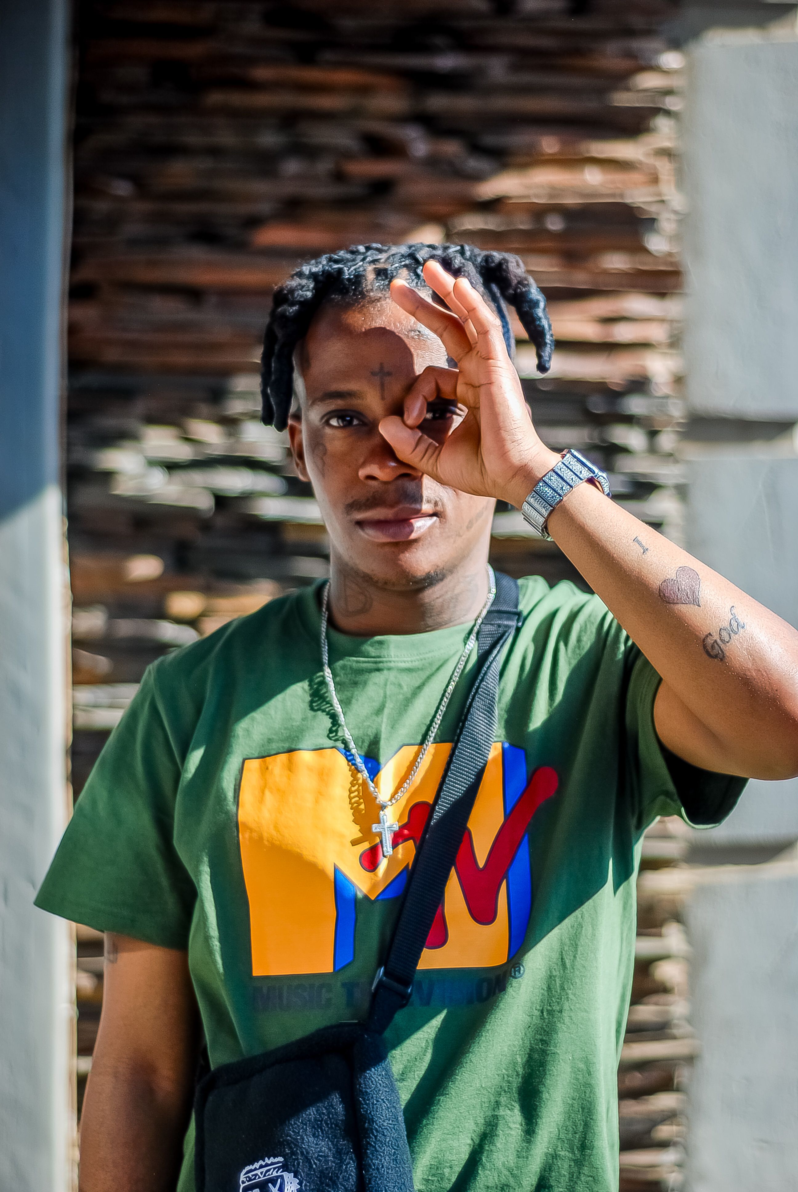 A young adult making a hand gesture outdoors, wearing a casual t-shirt and accessories.