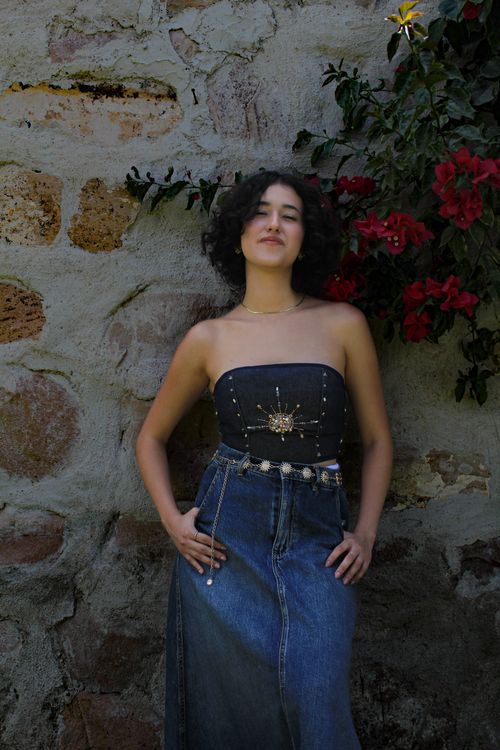 Free Confident woman in stylish denim and black top poses by a rustic stone wall adorned with flowers. Stock Photo