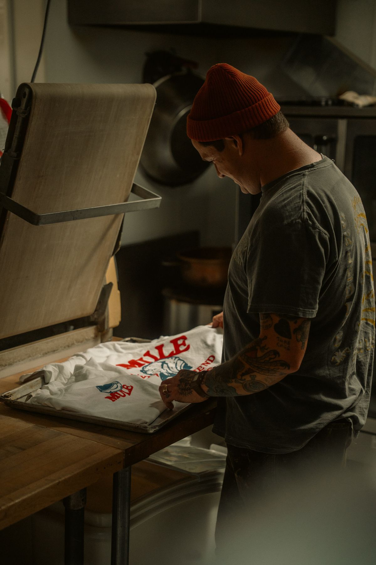 Printer in a red beanie laying a printed white shirt on a screen-printing press in a workshop