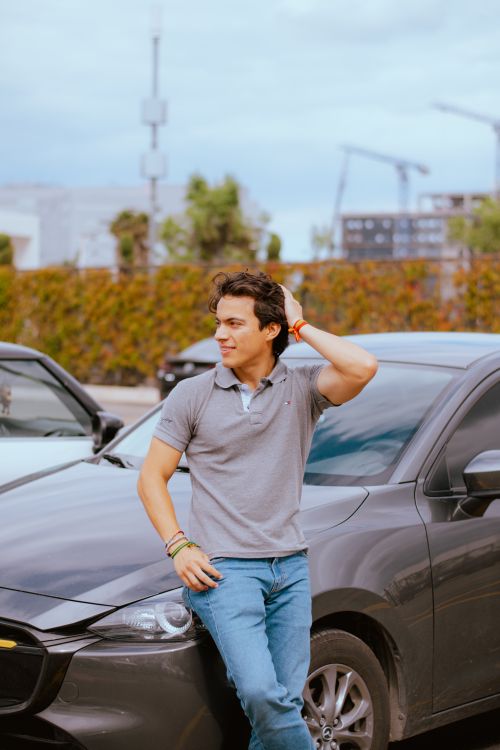 Free A young man in casual attire leans against a car outdoors, exuding confidence and style. Stock Photo
