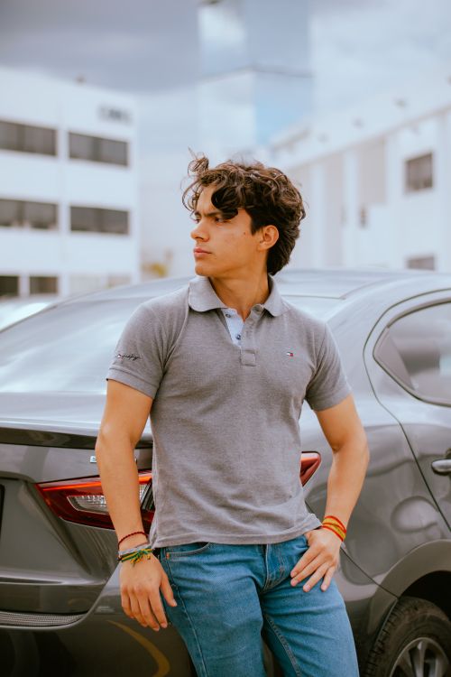 Free Young man in casual attire leaning on car with soft urban backdrop, displaying a relaxed expression. Stock Photo