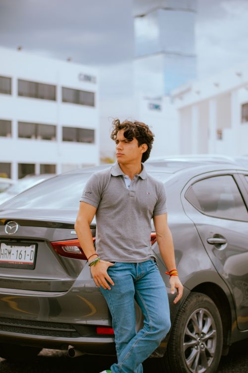 Free Fashionable young man in jeans and polo shirt leaning on car in cityscape setting. Stock Photo