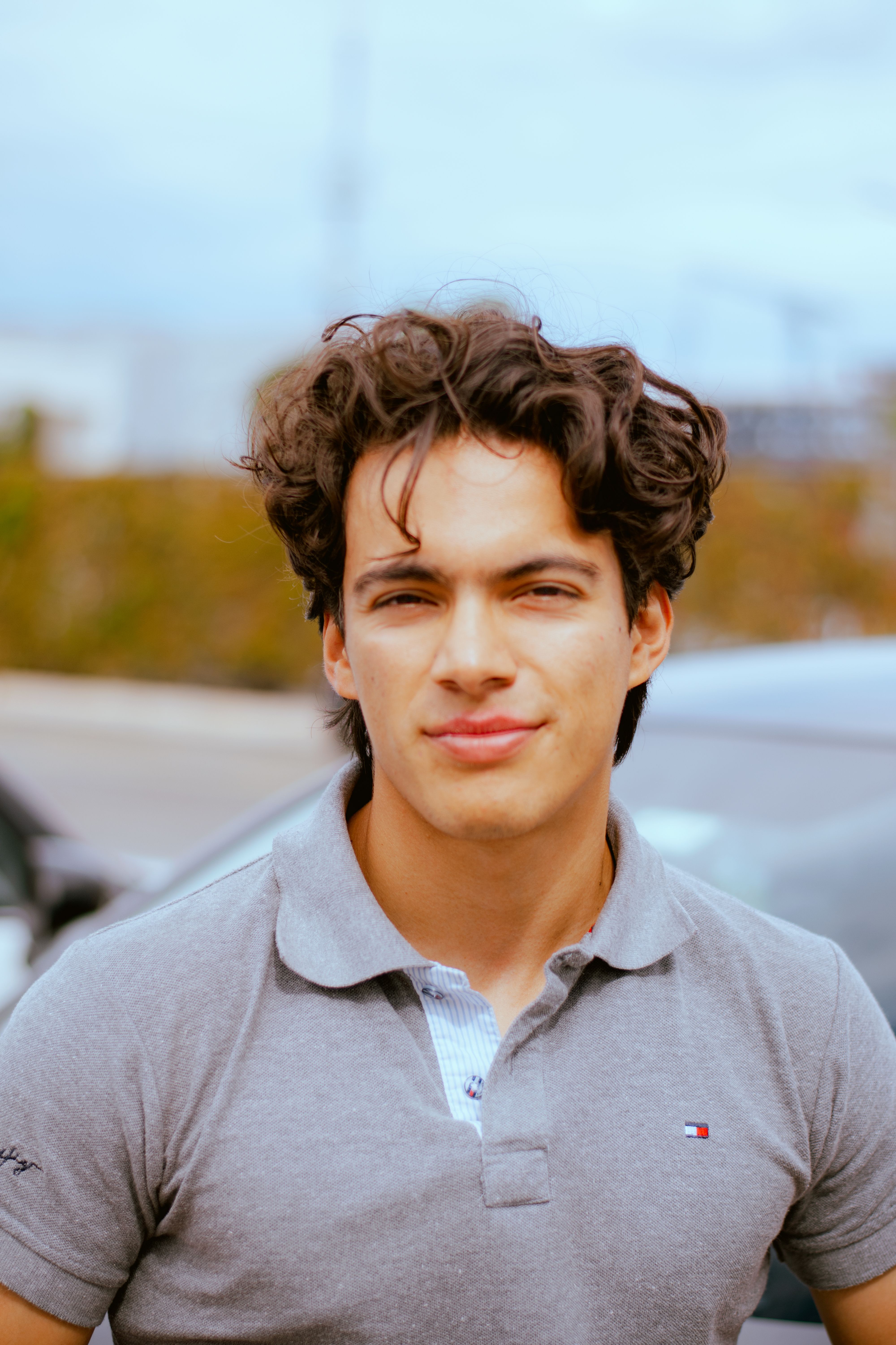 Portrait of a young curly-haired man outdoors, wearing a grey polo shirt with a soft expression.