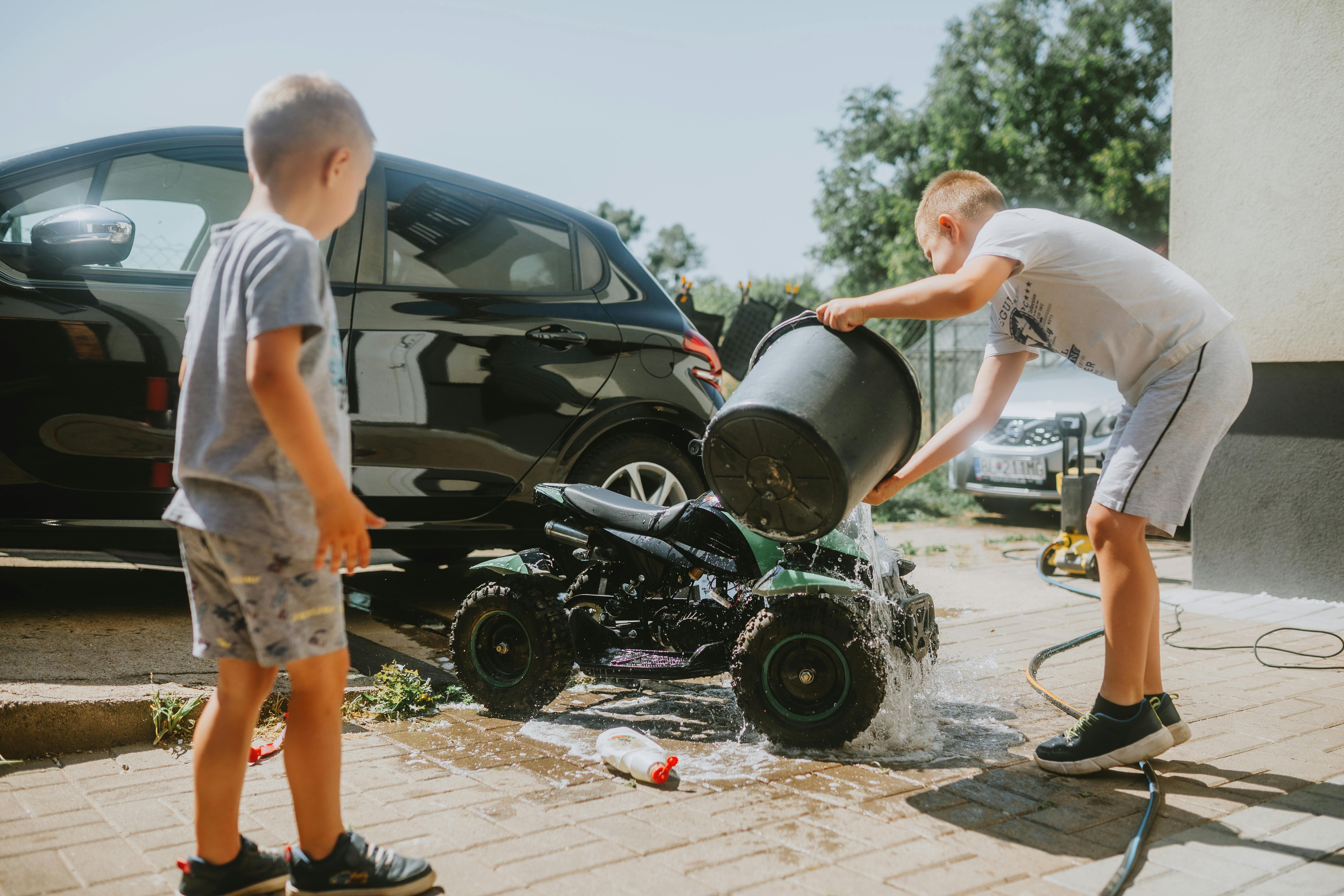Two boys wash a toy ATV with a bucket near a car in a sunny driveway.