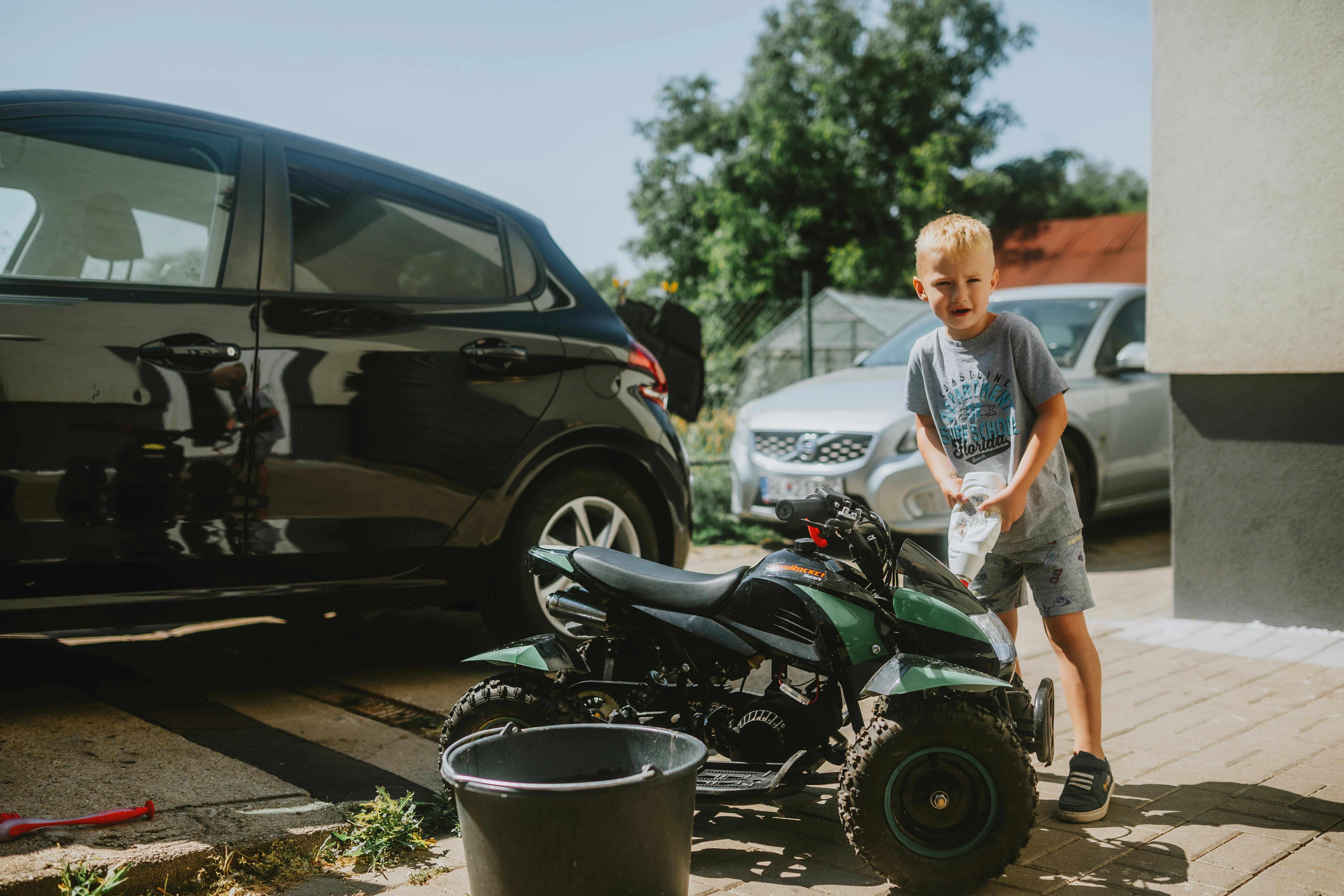 A young boy washes a toy ATV next to parked cars on a sunny day.