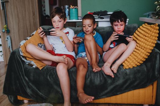 Three boys engrossed in mobile games while sitting on a couch at home, reflecting modern childhood leisure.