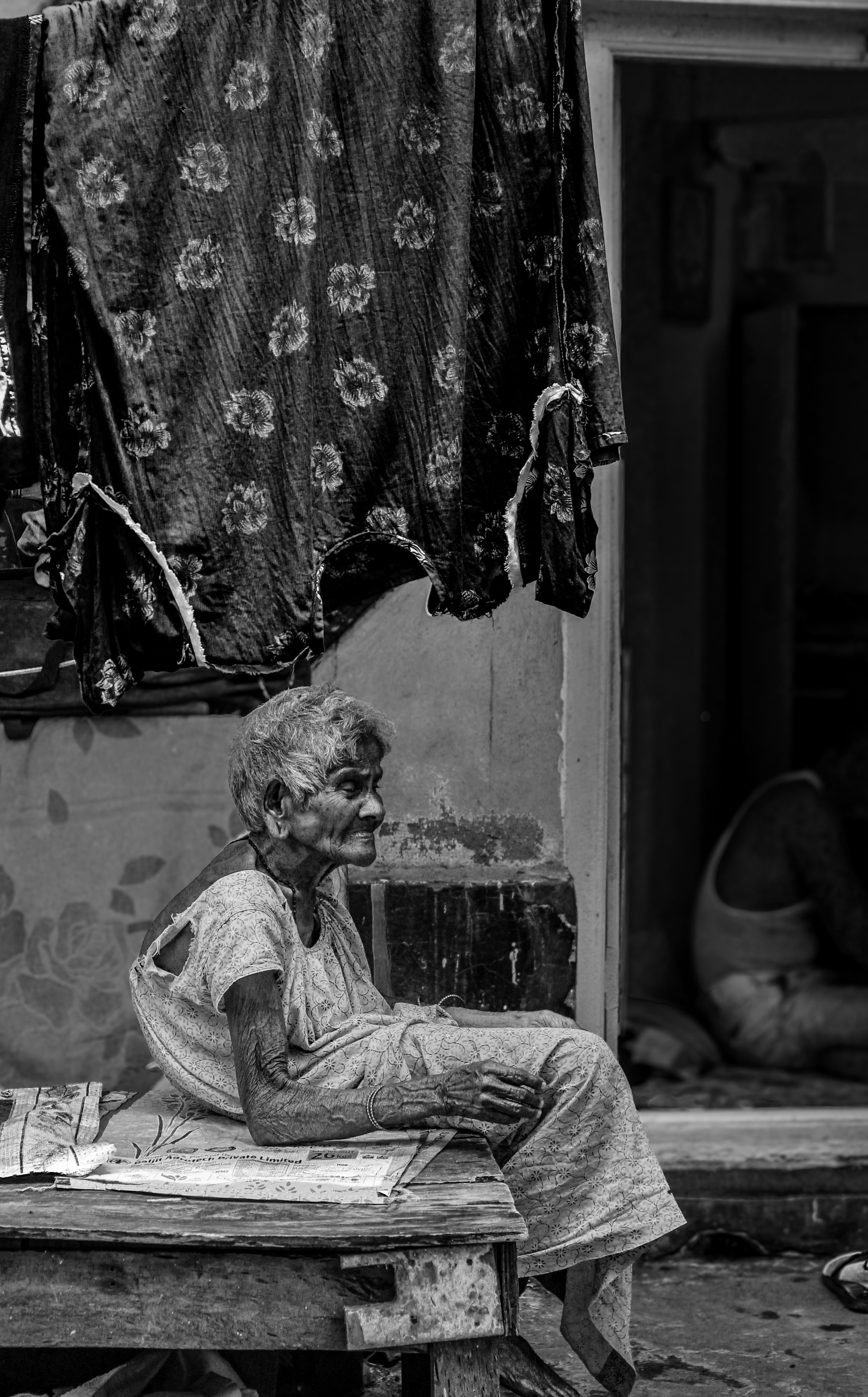 Fotomural Fotografía en blanco y negro de una anciana sentada afuera de una casa rústica, evocando nostalgia.