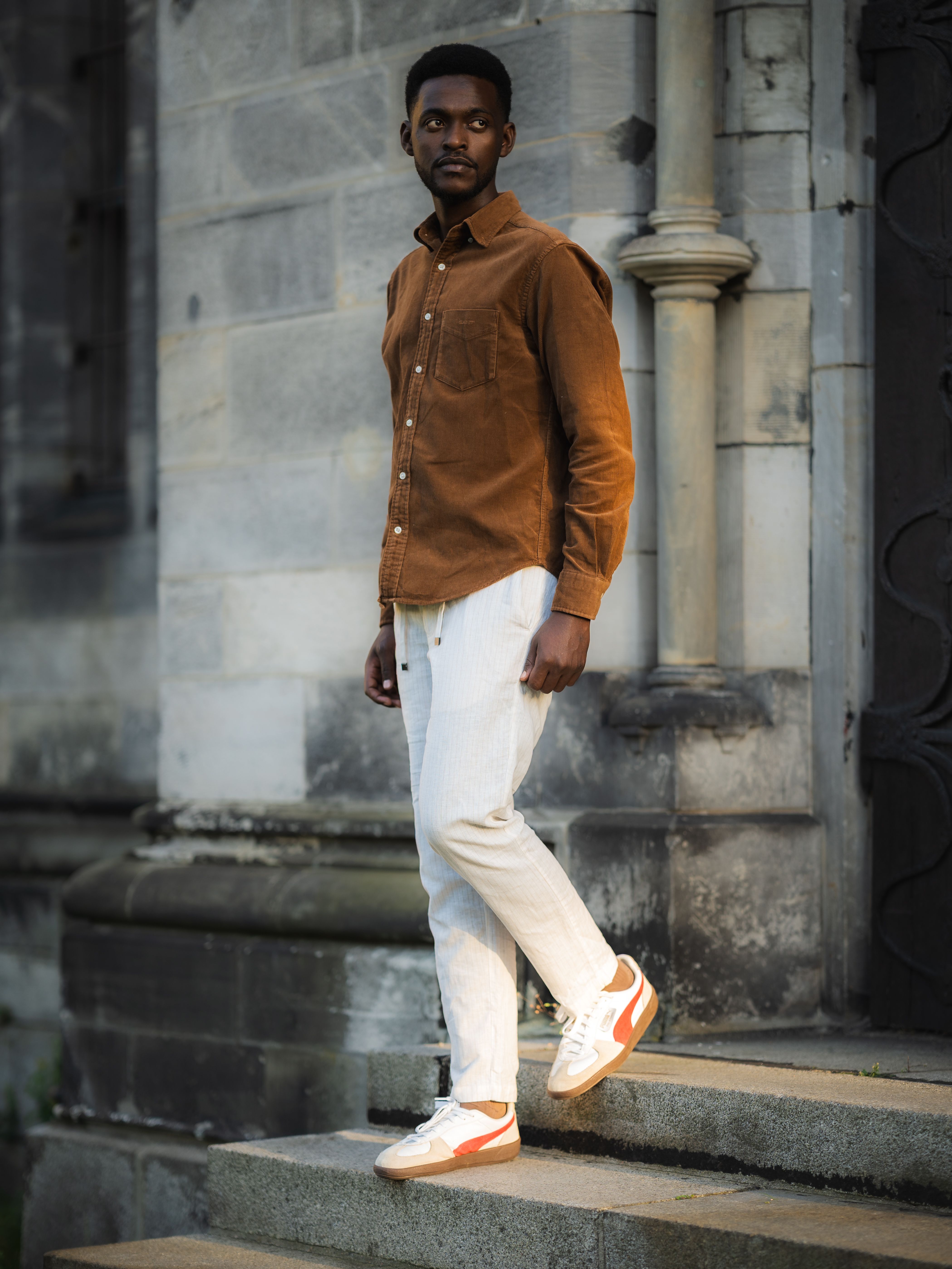 Fashionable man in a brown shirt and white pants, posing on urban steps with an elegant backdrop.
