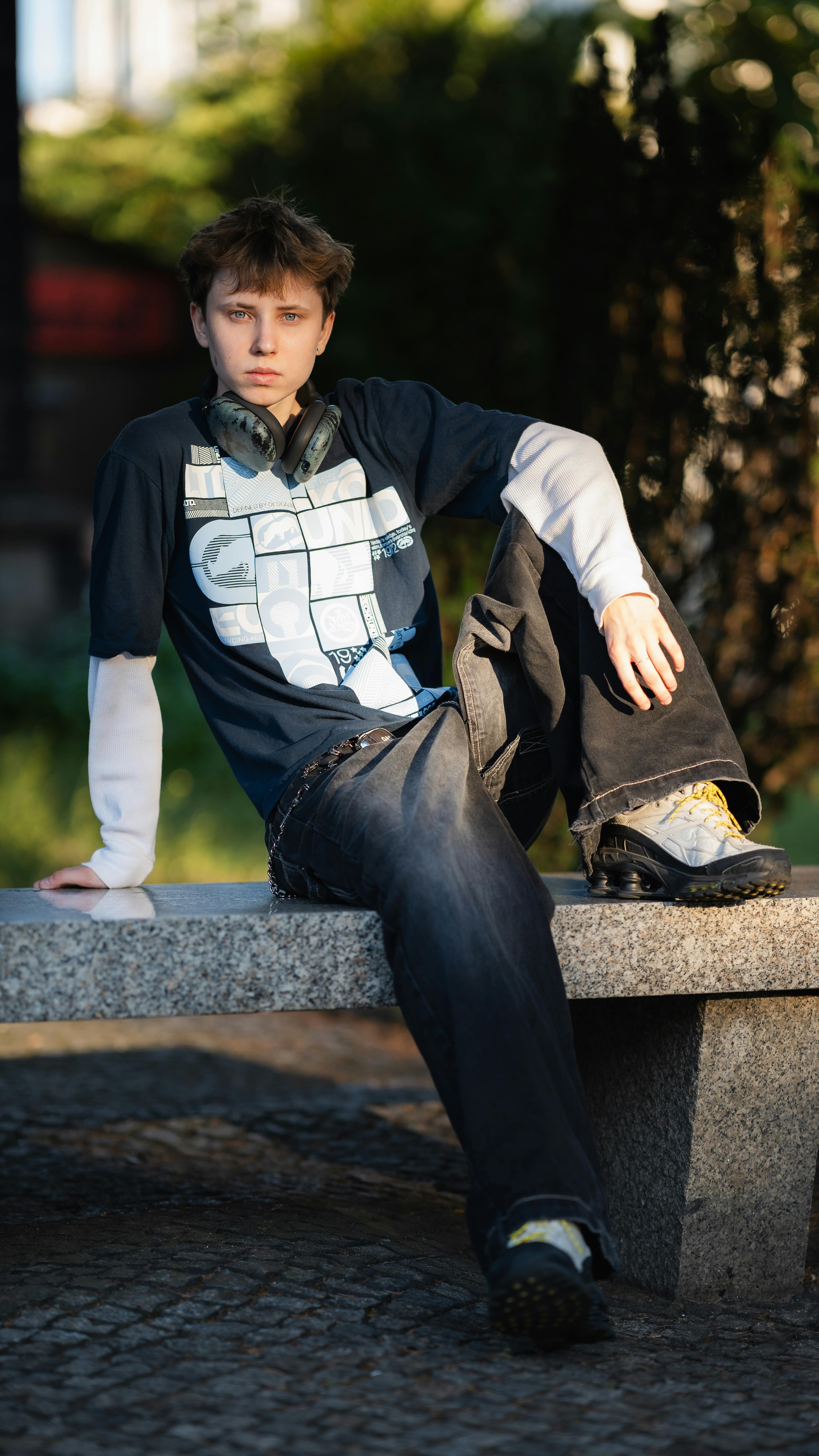 Gratis Adolescente vestido con ropa informal sentado en un banco de piedra al aire libre con auriculares alrededor del cuello. Foto de stock