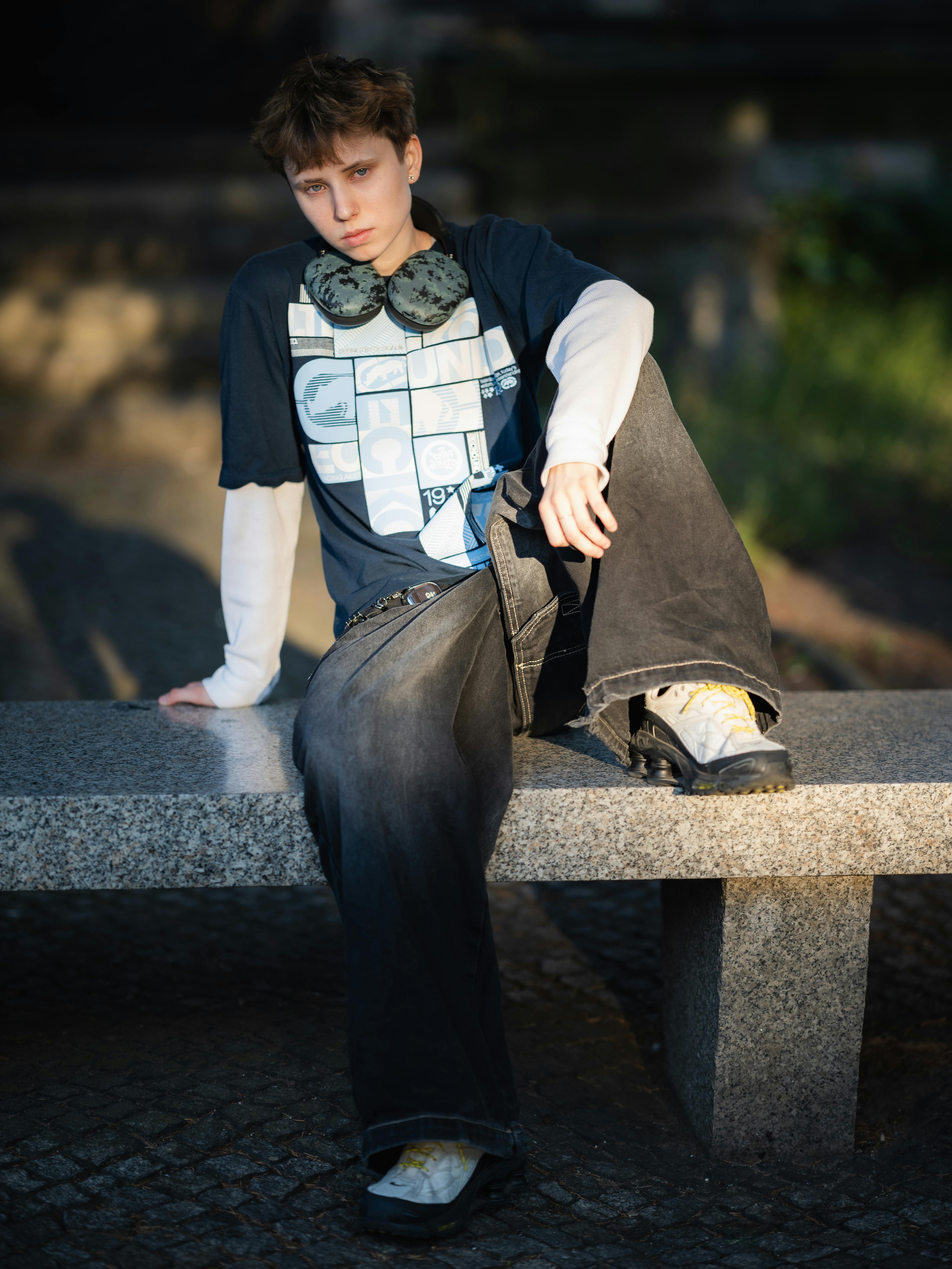 Kostenlos Teenager in stylischem Outfit mit Kopfhörern sitzt auf einer Steinbank und genießt einen sonnigen Tag im Freien. Stock-Foto