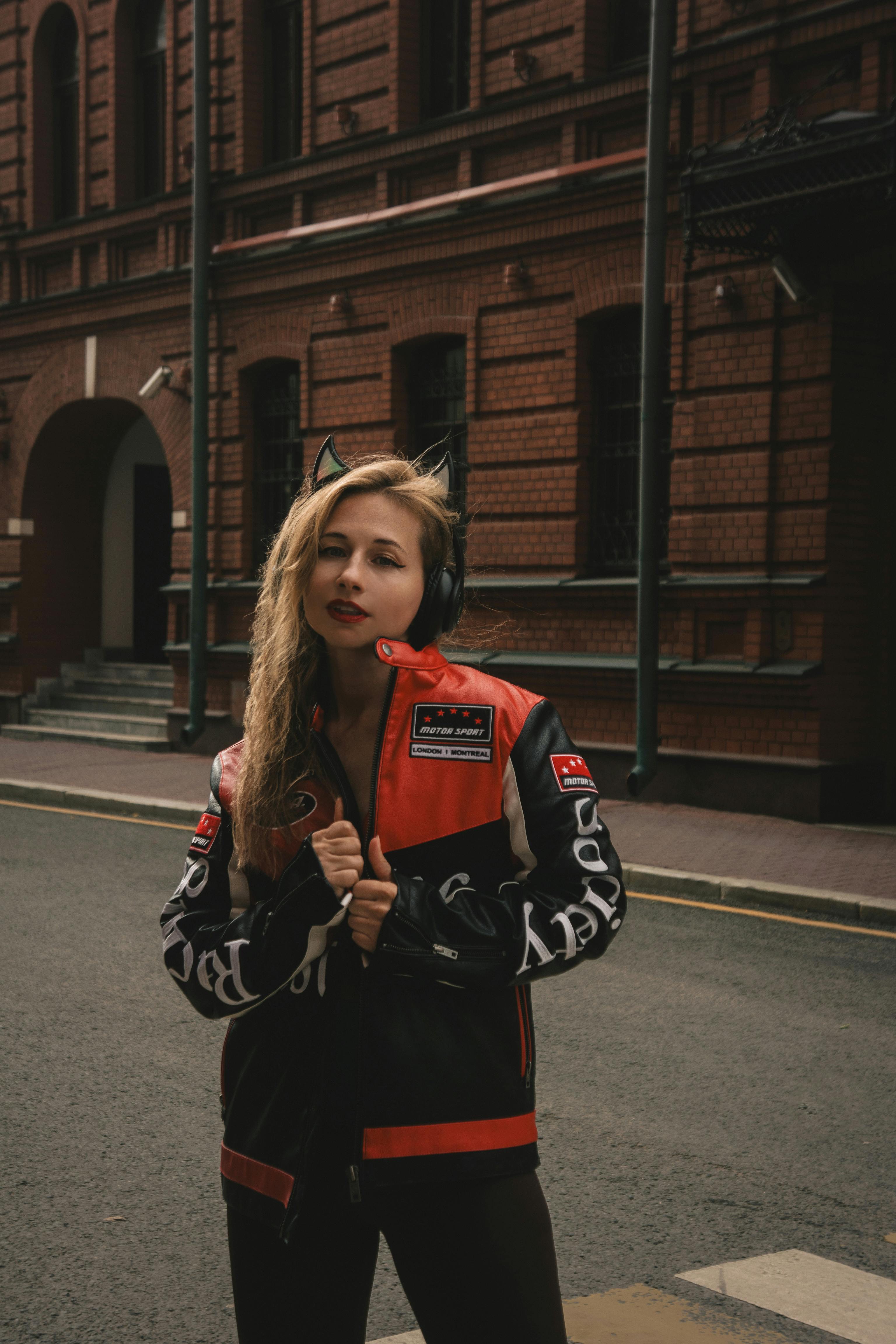Stylish woman with headphones stands confidently in city street with brick architecture.