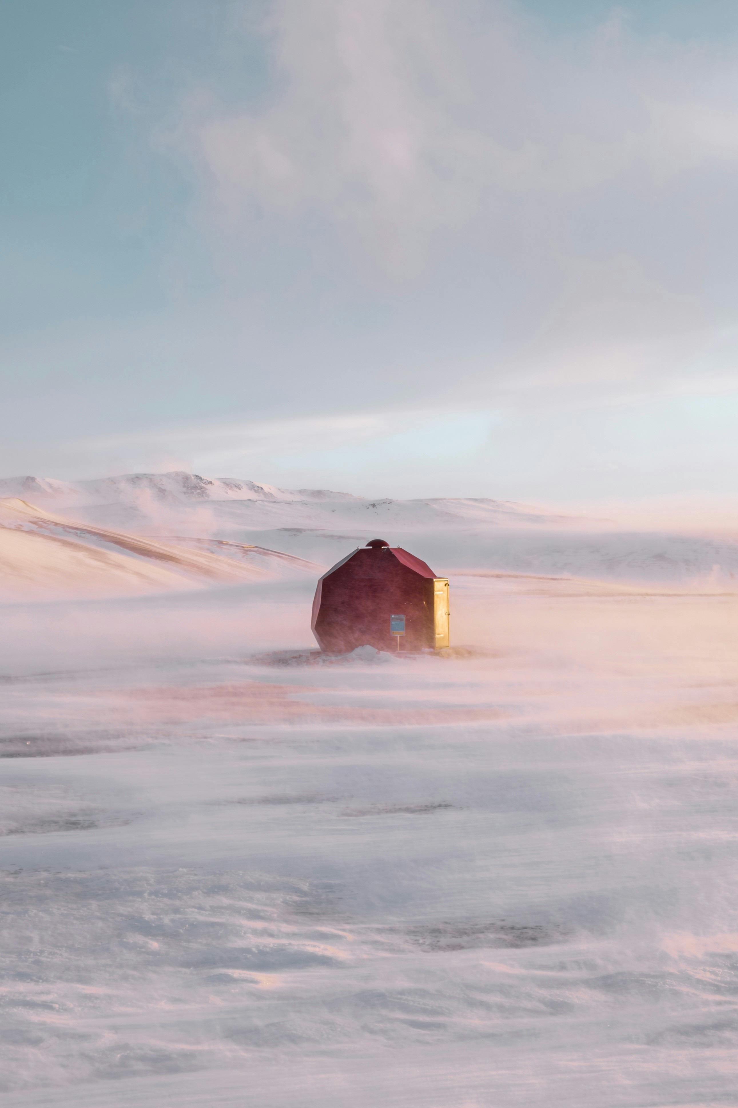 A red cabin stands solitary on a snowy landscape during sunrise, enveloped by soft pastel skies.