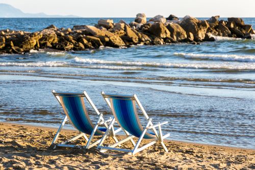 Free Two empty beach chairs facing the ocean with gentle waves and a rocky barrier in the background. Stock Photo