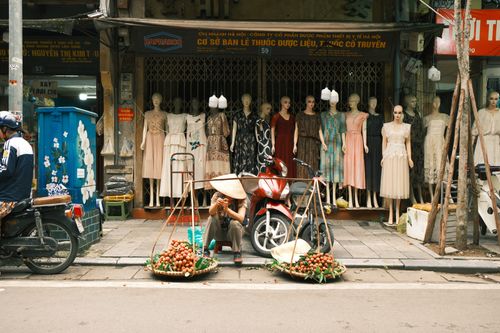 Free Street scene in Hanoi featuring a vendor selling fruits in front of a mannequin display. Stock Photo