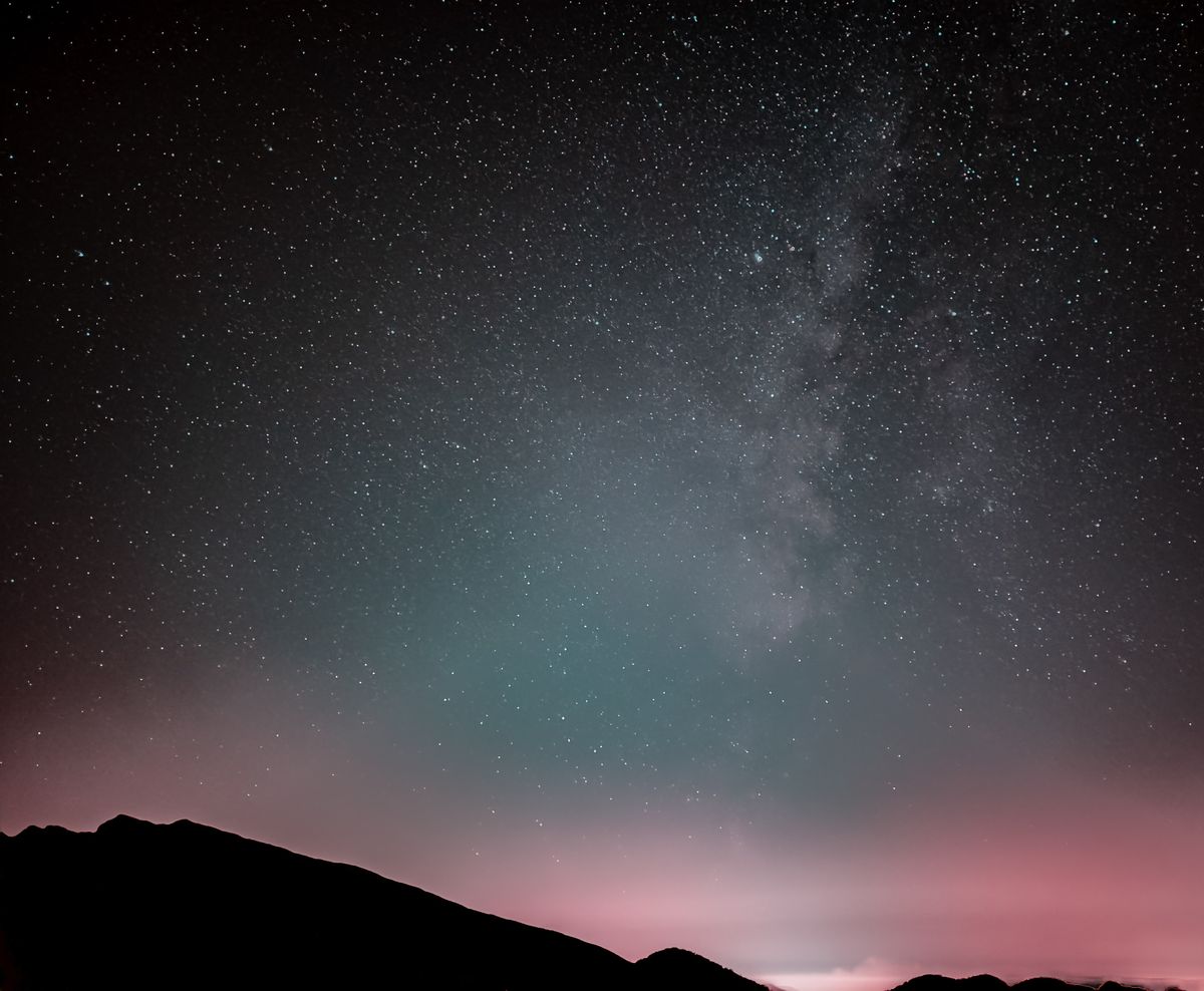 The Milky Way over a dark mountain silhouette, a faint pink-green airglow band low on the horizon, stars uncountable by eye.