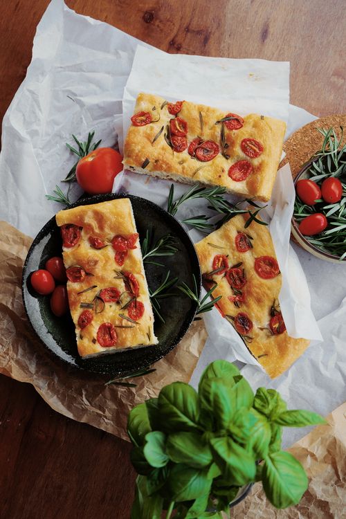 Free Delicious focaccia topped with cherry tomatoes and rosemary, styled with fresh herbs. Stock Photo