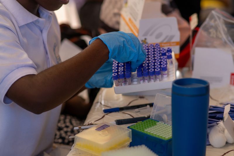 A gloved health worker sorts labeled blood-collection tubes at a testing table.