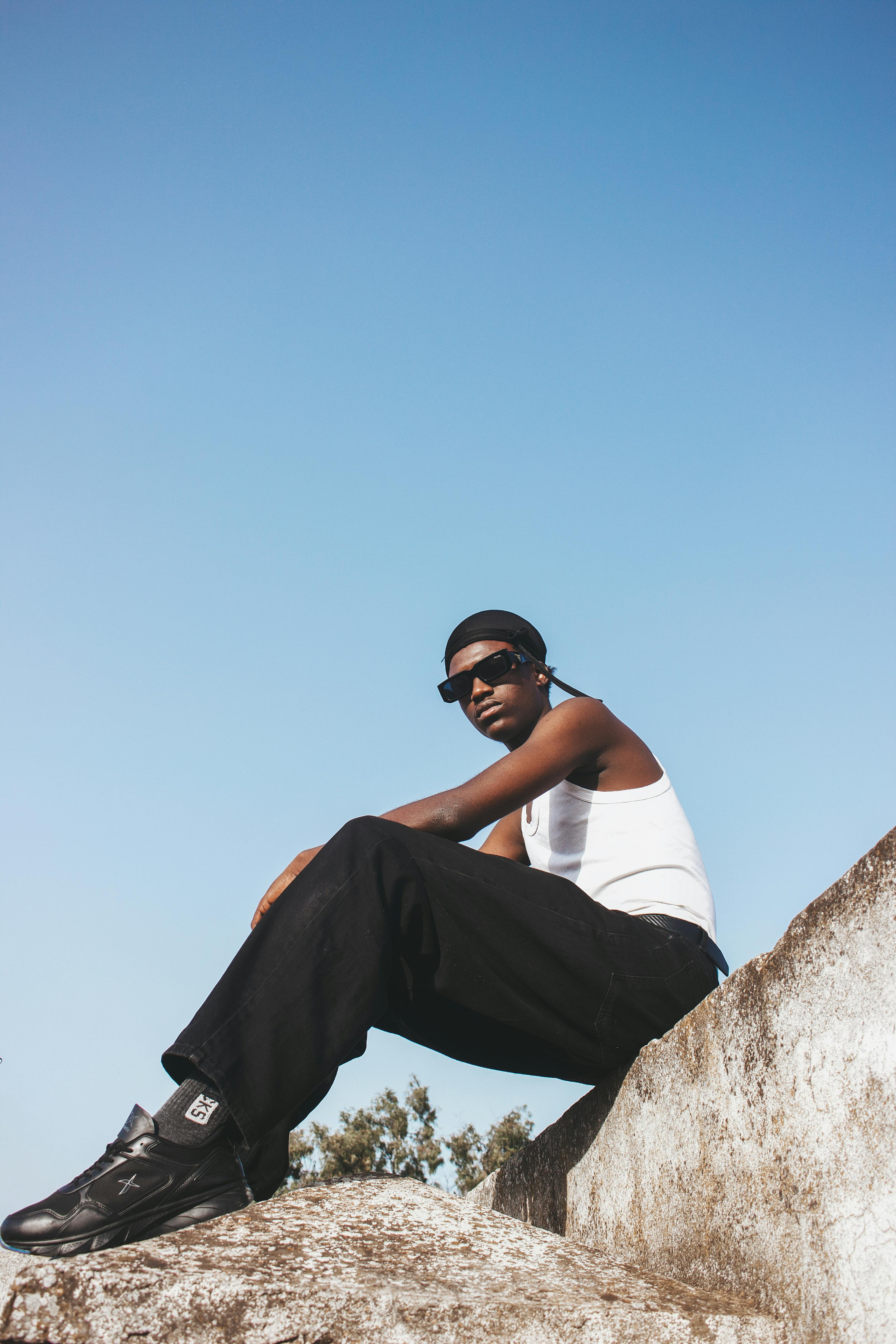 A young man in sunglasses and casual attire sitting on a rock against a clear blue sky.