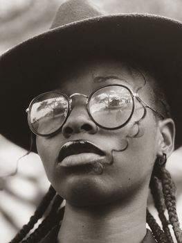 Black and white portrait of a woman wearing a hat and glasses, with a moody expression.