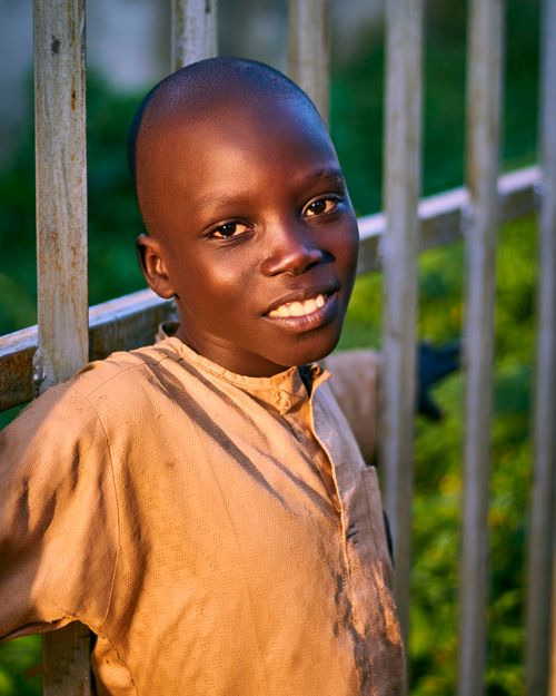 Free A smiling child leaning against a metal fence with greenery in the background. Stock Photo