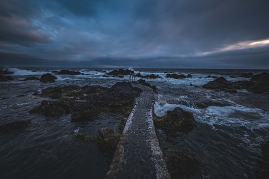 A moody seascape with a stone pathway leading into the ocean under a dramatic twilight sky.