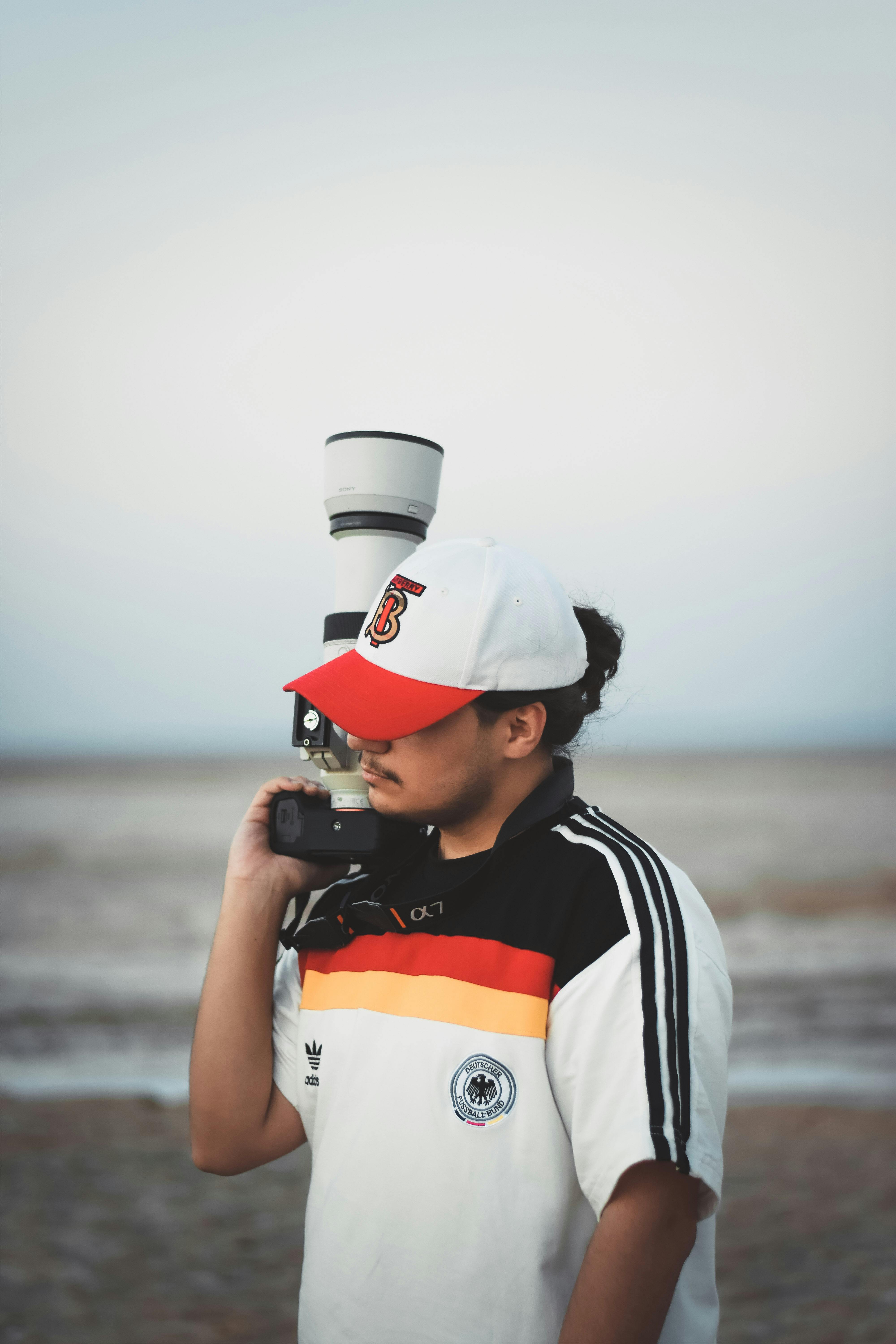 A young man in an adidas jersey and cap holds a camera outdoors, showcasing casual photography style.