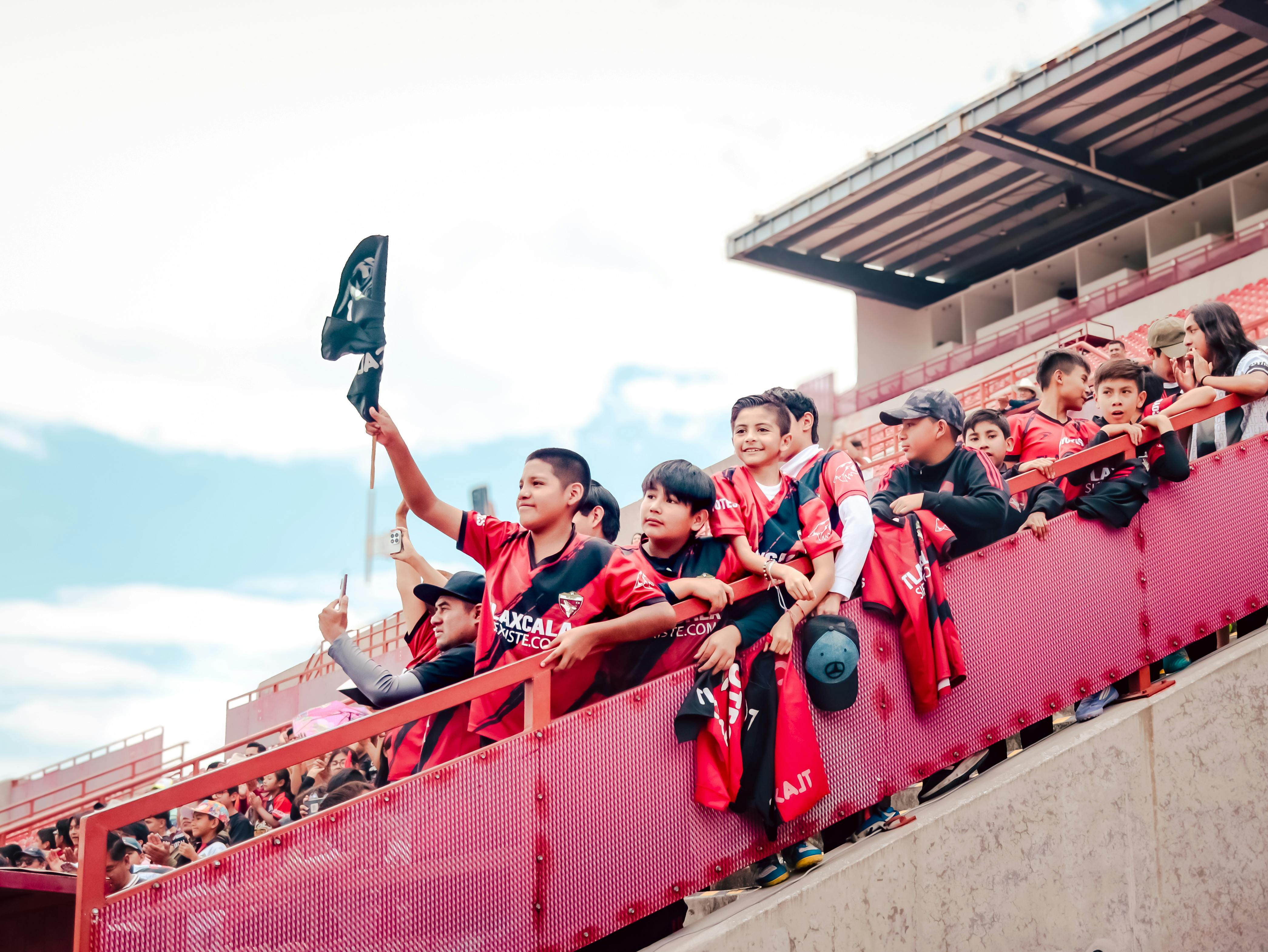 A group of enthusiastic young soccer fans cheering in the stands during a daytime football match.