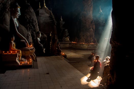 A monk meditates in a dimly lit cave temple with Buddha statues and candle light.