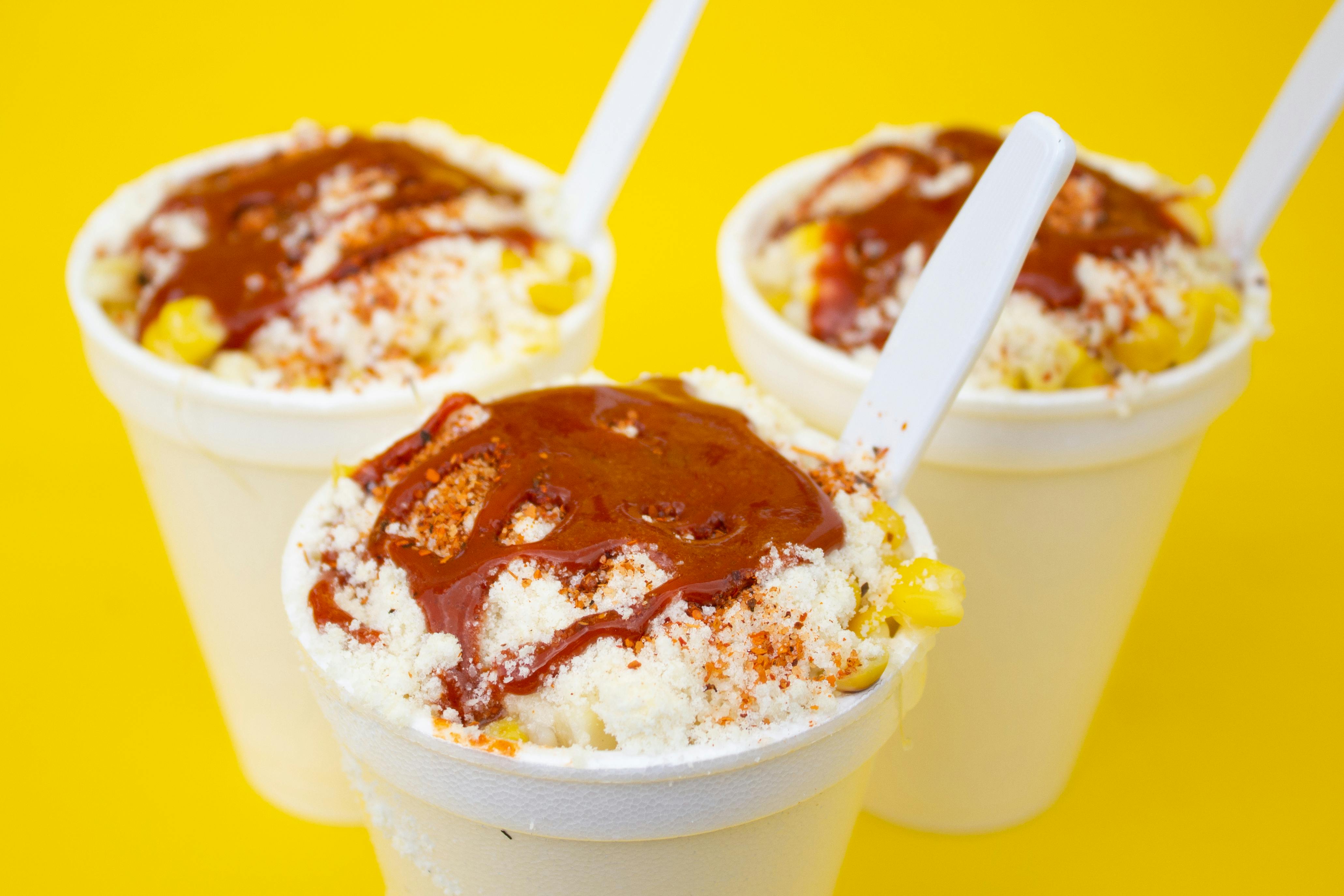 Colorful and appetizing Mexican elotes in white cups with toppings, captured on a vibrant yellow backdrop.