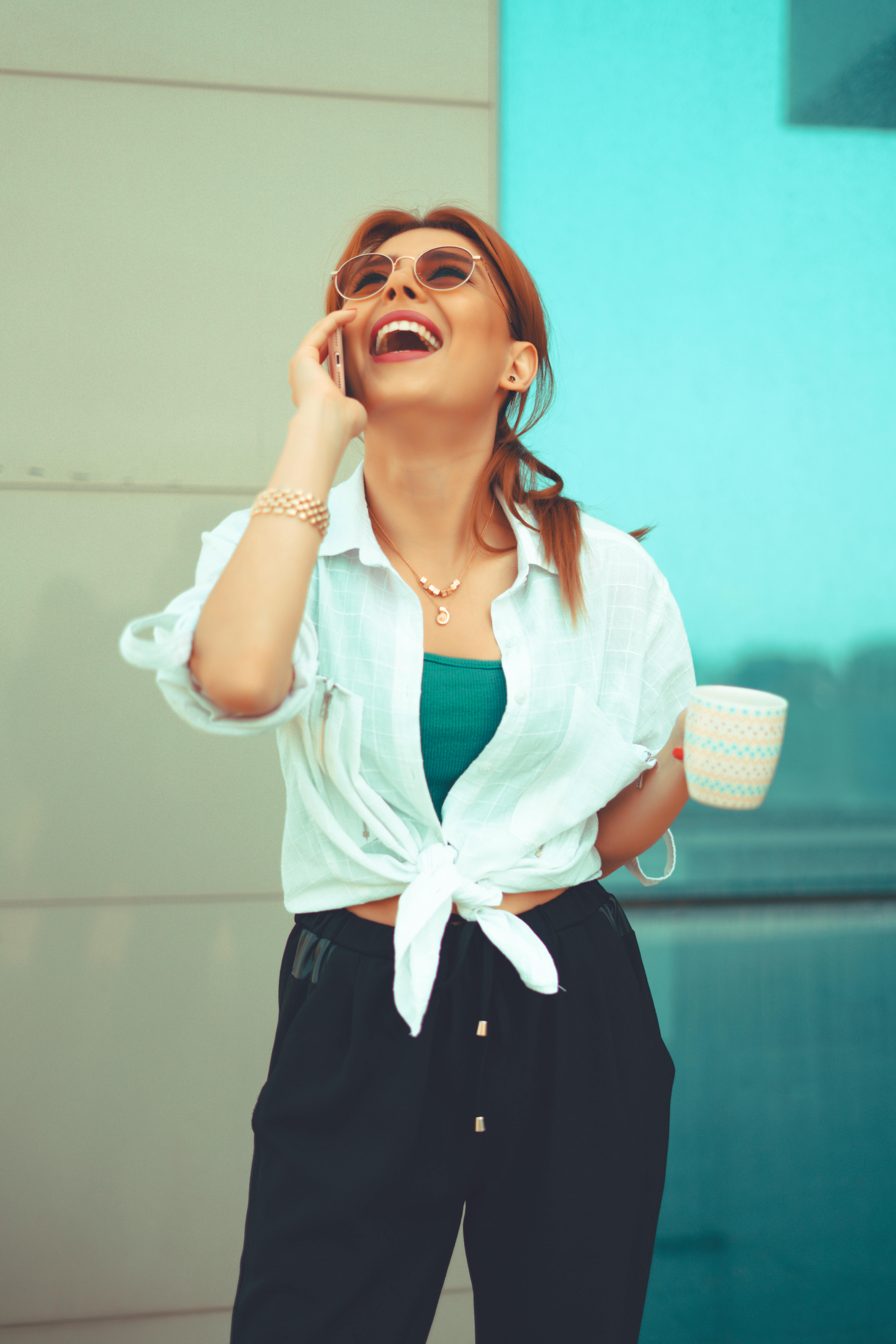 A young woman laughs while talking on the phone outdoors, holding a mug.