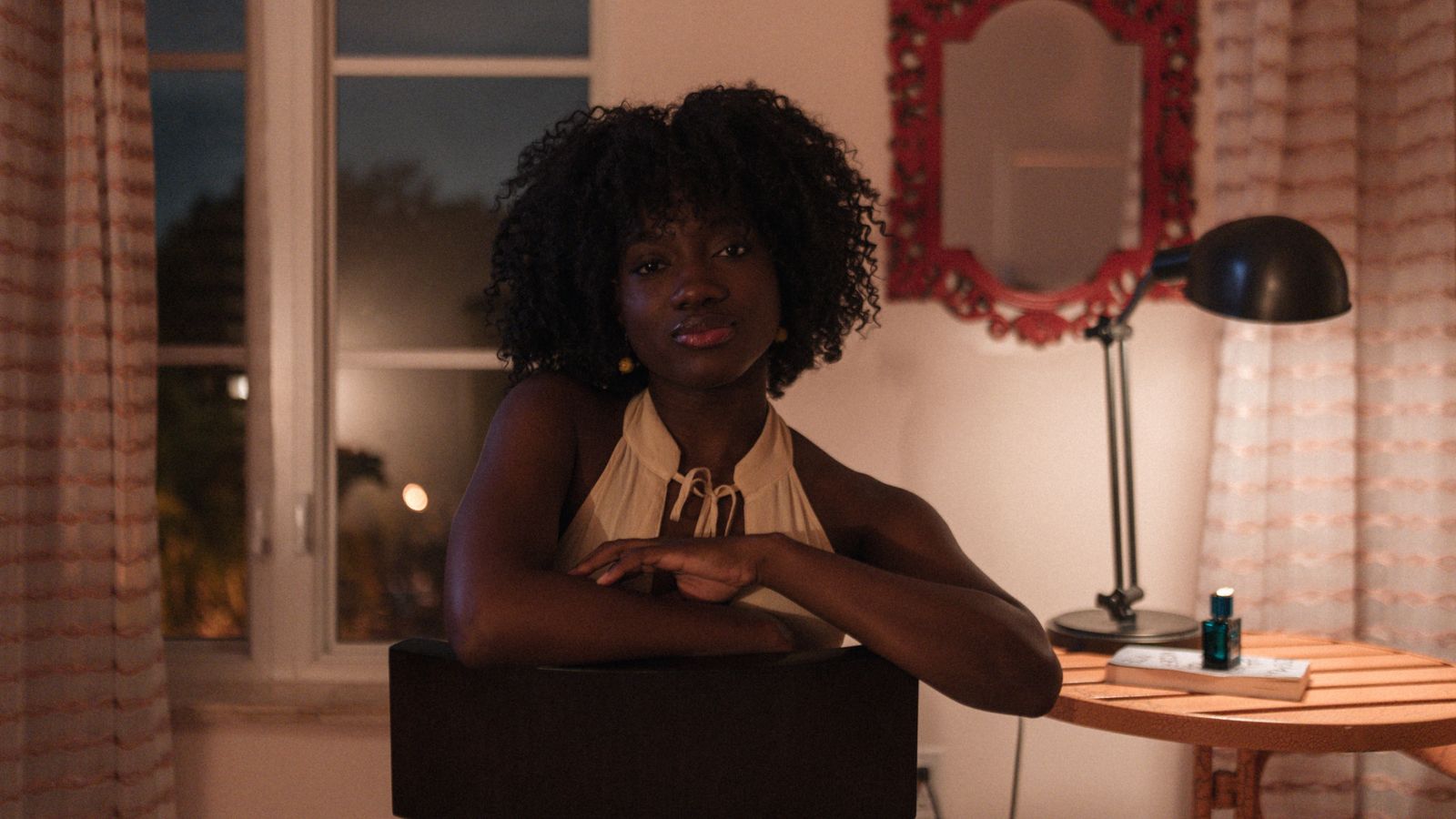 Woman with natural curls leans on a chair at home beside a warm lamp and small blue bottle