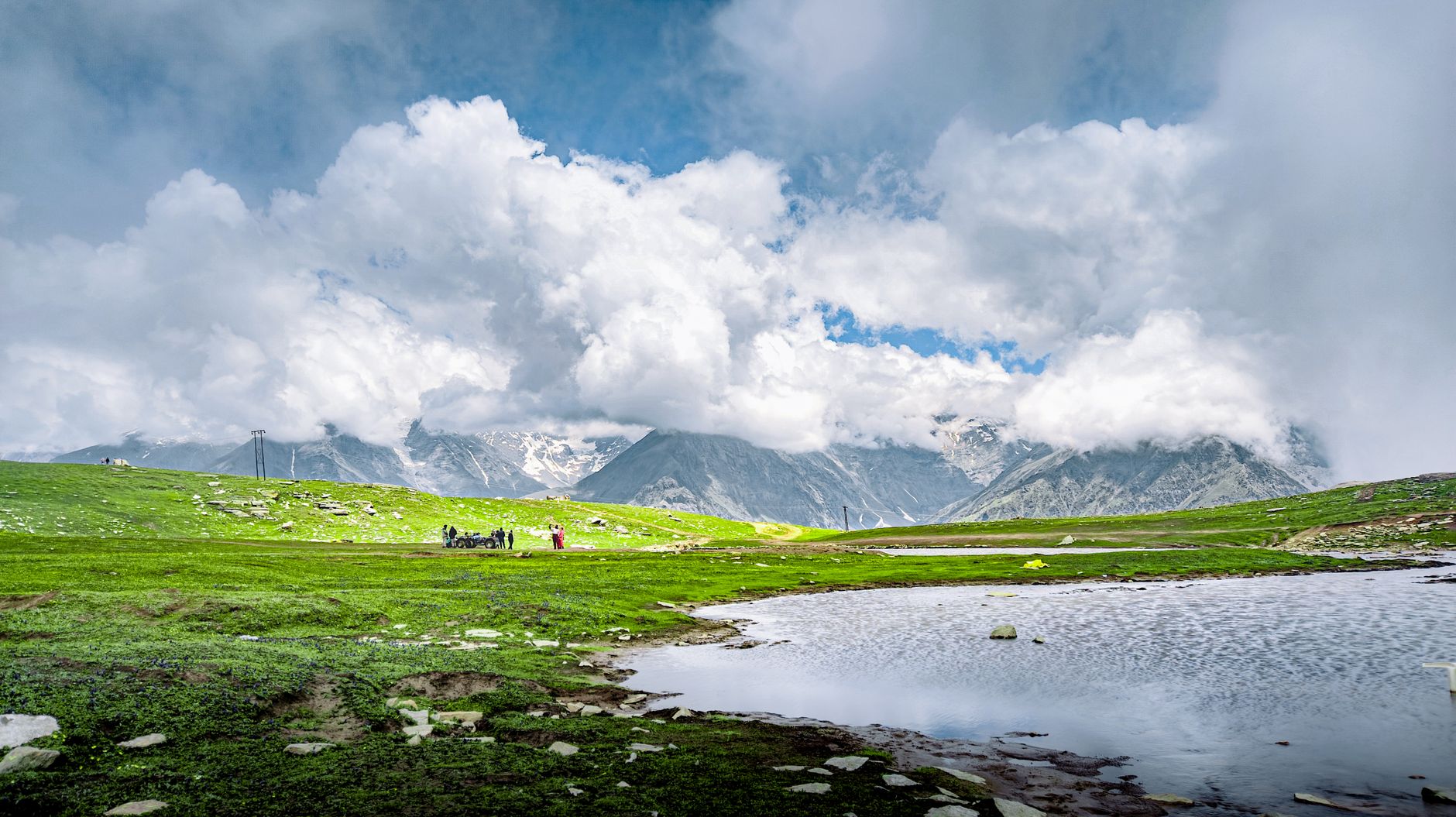A breathtaking view of the Himalayas with lush green fields, a pristine lake, and dramatic clouds overhead.