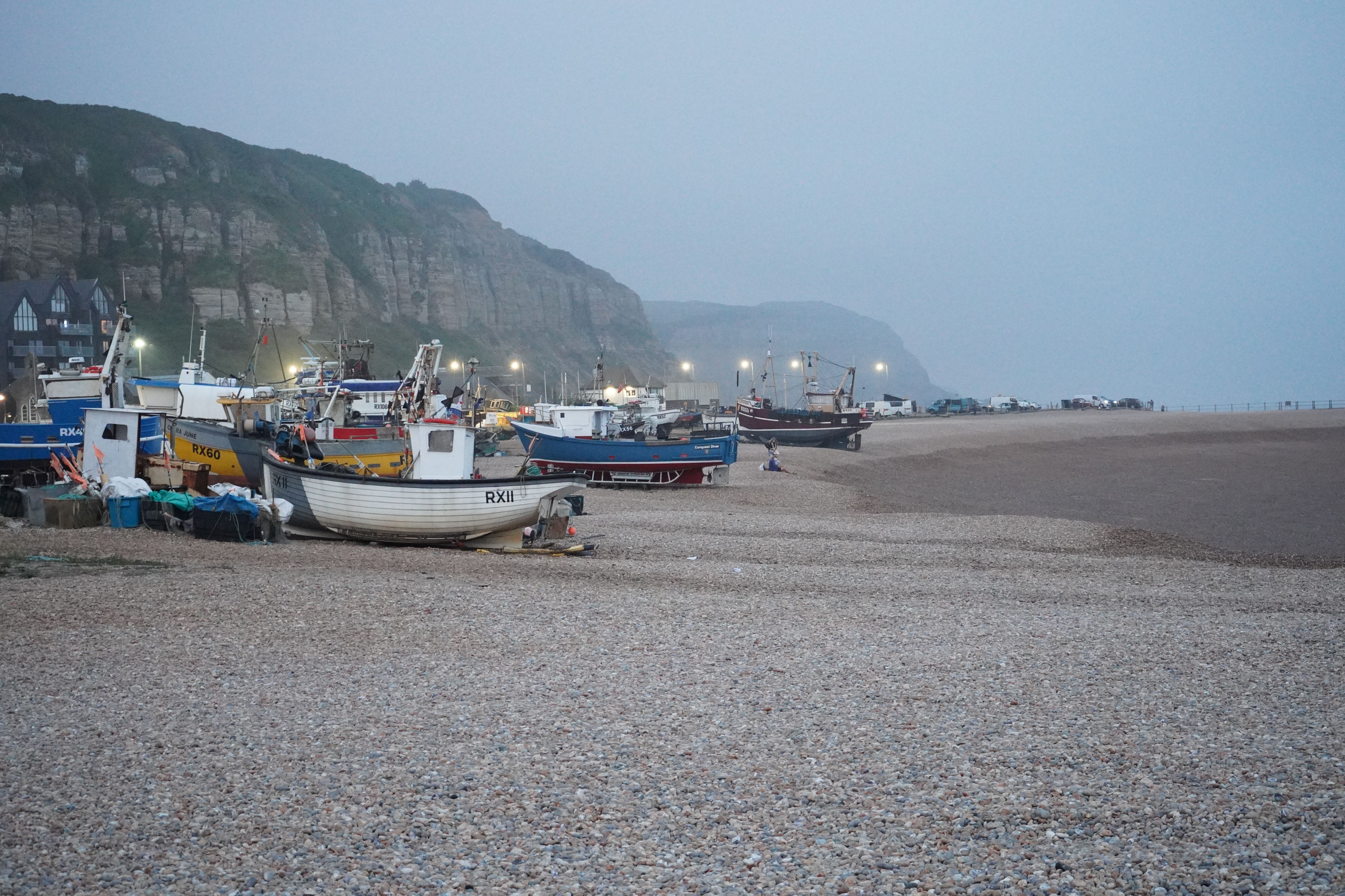 A serene scene of fishing boats at dusk on a pebble beach with cliffs in the background.