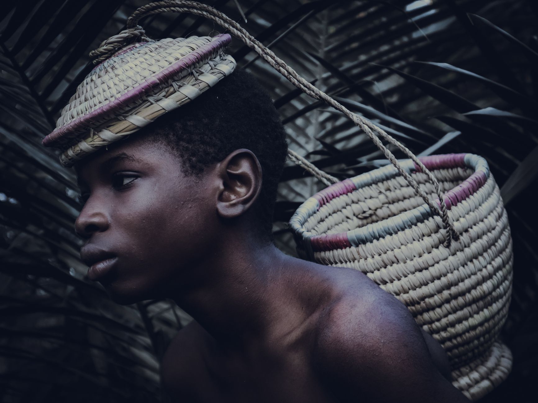 A contemplative young boy carries a woven basket on his head in a natural setting, Abuja, Nigeria.