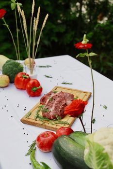 Outdoor setting with fresh vegetables and raw meat on a wooden board garnished with herbs.