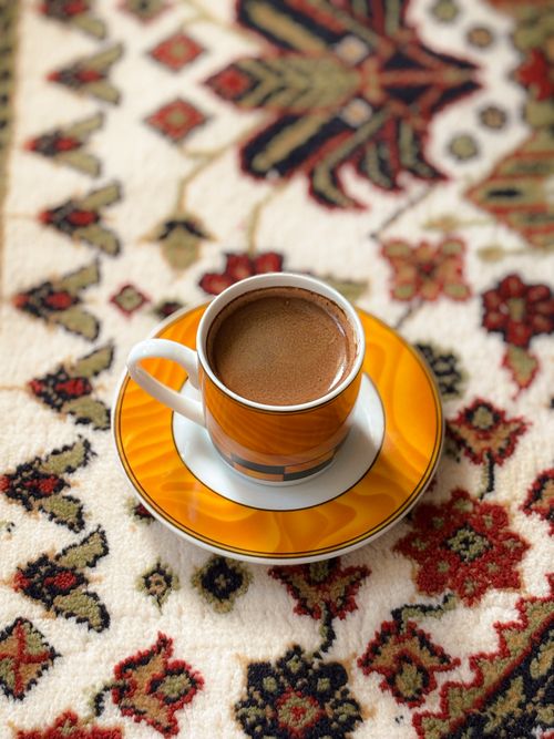 Free Close-up of a coffee cup with frothy top on a colorful patterned rug, creating a warm and inviting scene. Stock Photo