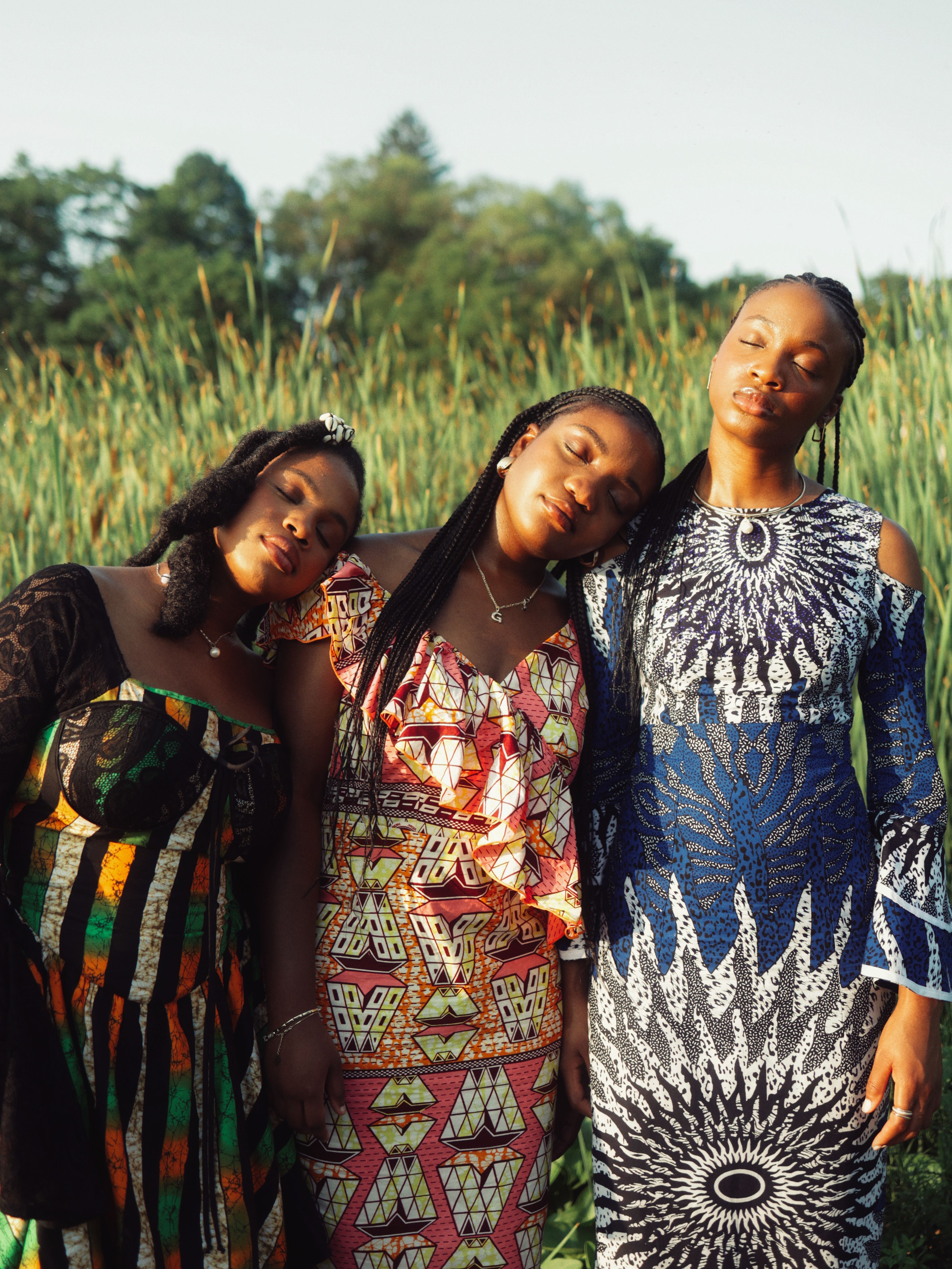 Three women in vibrant African dresses peacefully standing together with eyes closed in a serene outdoor setting.