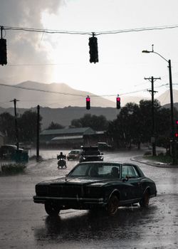 Vintage car drives through a rain-soaked intersection under gloomy sky with mountains in the distance.
