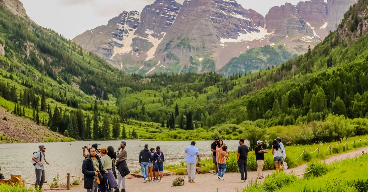 Visitors gather at Maroon Lake with Maroon Bells view, surrounded by lush summer greenery.